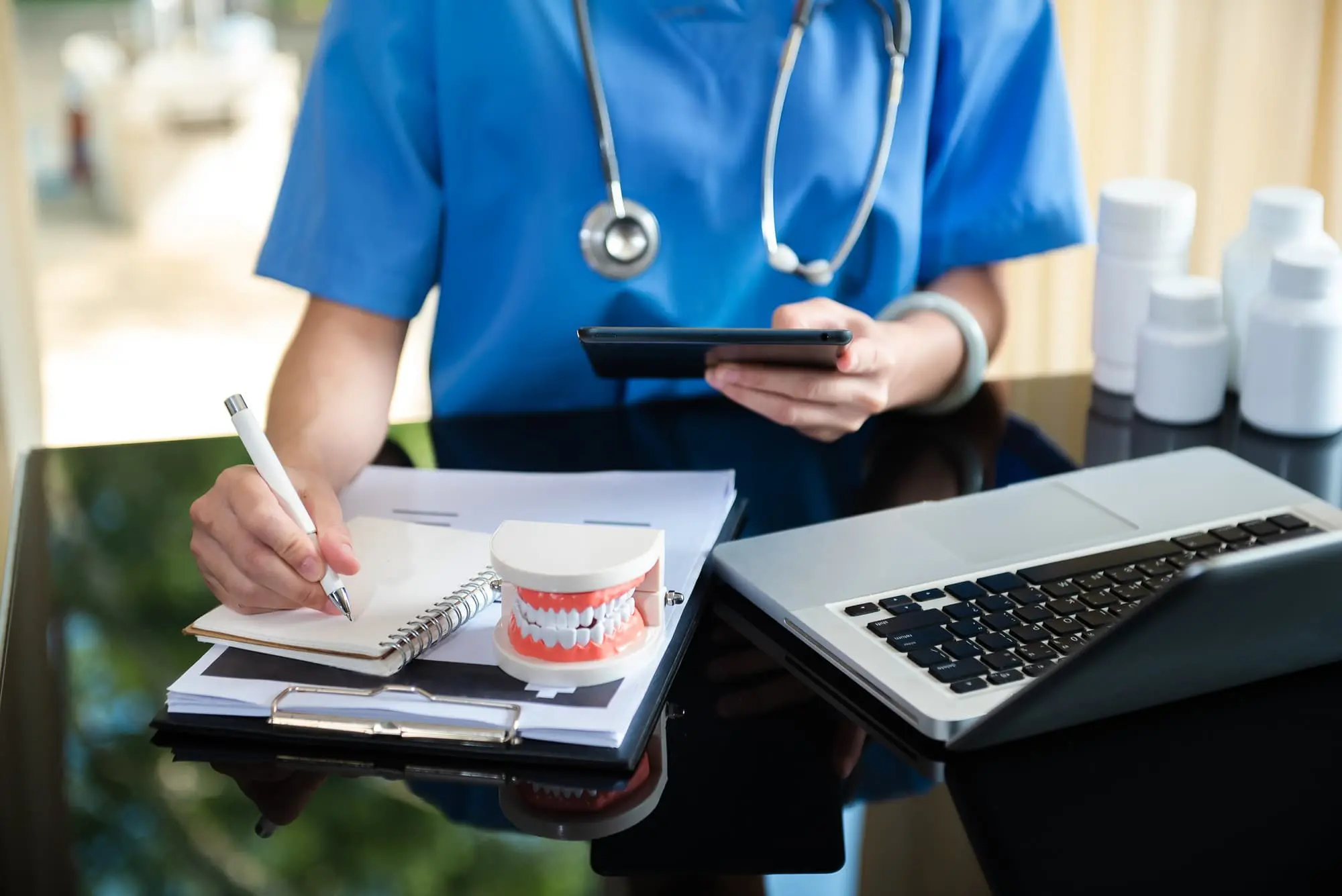 At Levine Orthodontics in Fort Myers, Cape Coral or Lehigh Acres, FL, someone in blue scrubs writes on a notepad and balances a phone amidst a dental model, laptop and medication bottles—likely reviewing orthodontic insurance plans.