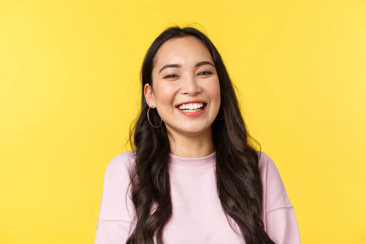 A woman with long dark hair, hoop earrings, and a light pink sweater—showing a warm smile with a slight underbite—appears against a plain yellow background at Levine Orthodontics in Fort Myers, Cape Coral or Lehigh Acres, FL.