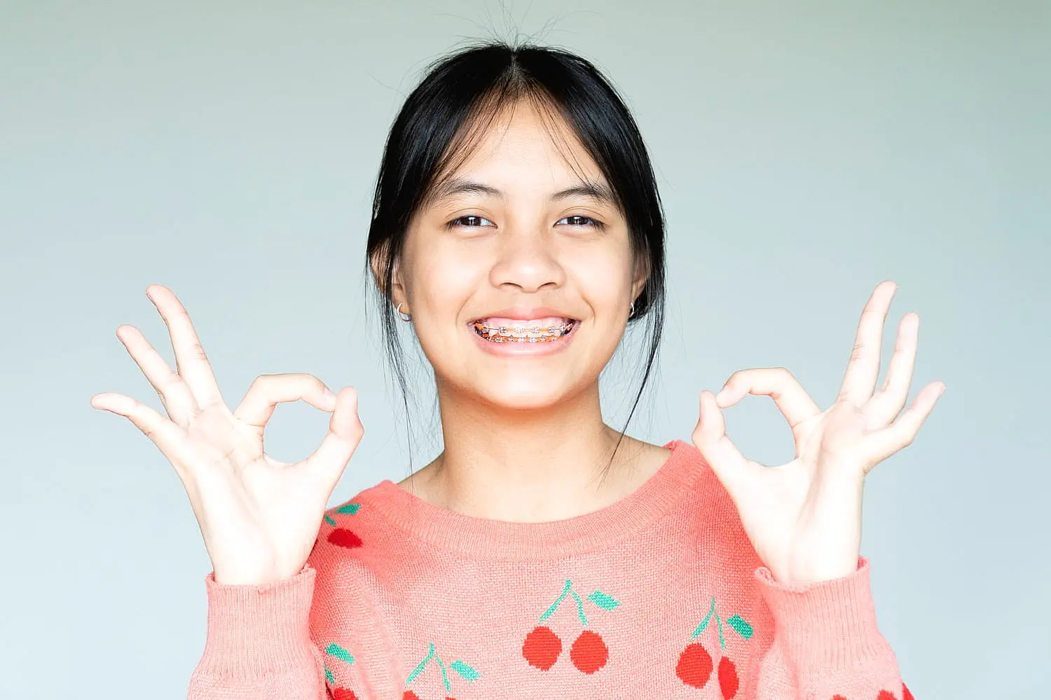 A smiling young person with metal braces, dressed in a pink sweater, holds up both hands making an "OK" gesture against a light background—representing Levine Orthodontics in Fort Myers, Cape Coral or Lehigh Acres, FL.