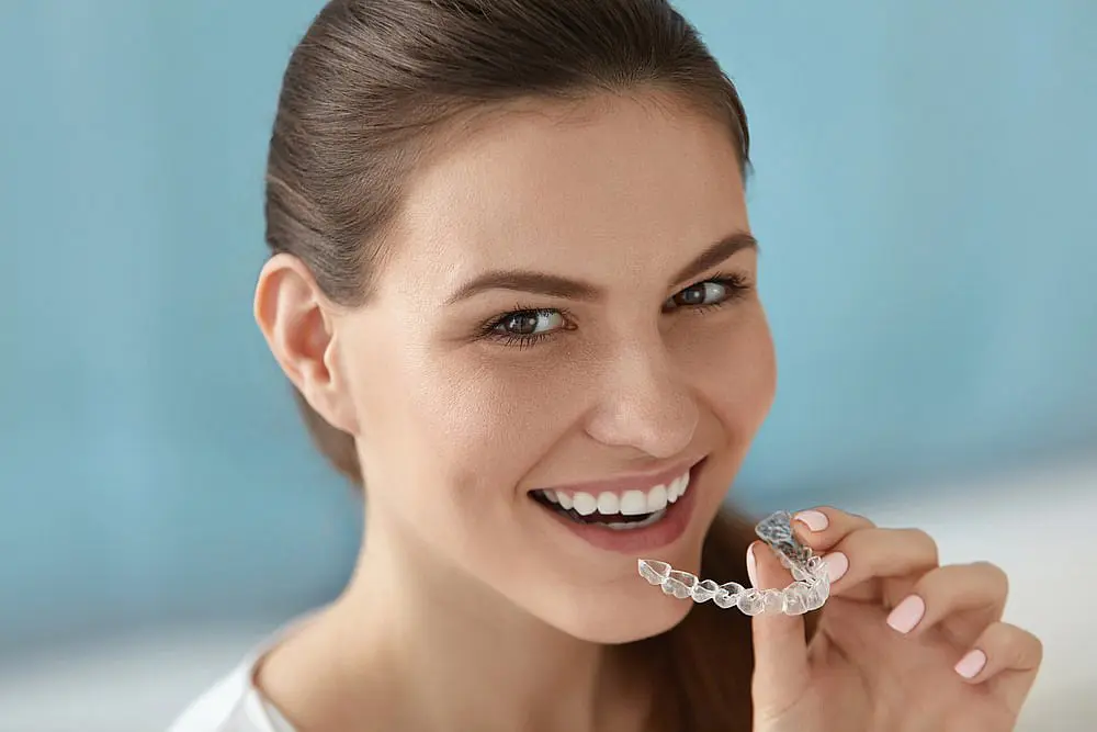 A woman smiles while holding Invisalign® Clear Aligners in front of her mouth against a light blue background at Levine Orthodontics in Lehigh Acres, FL.