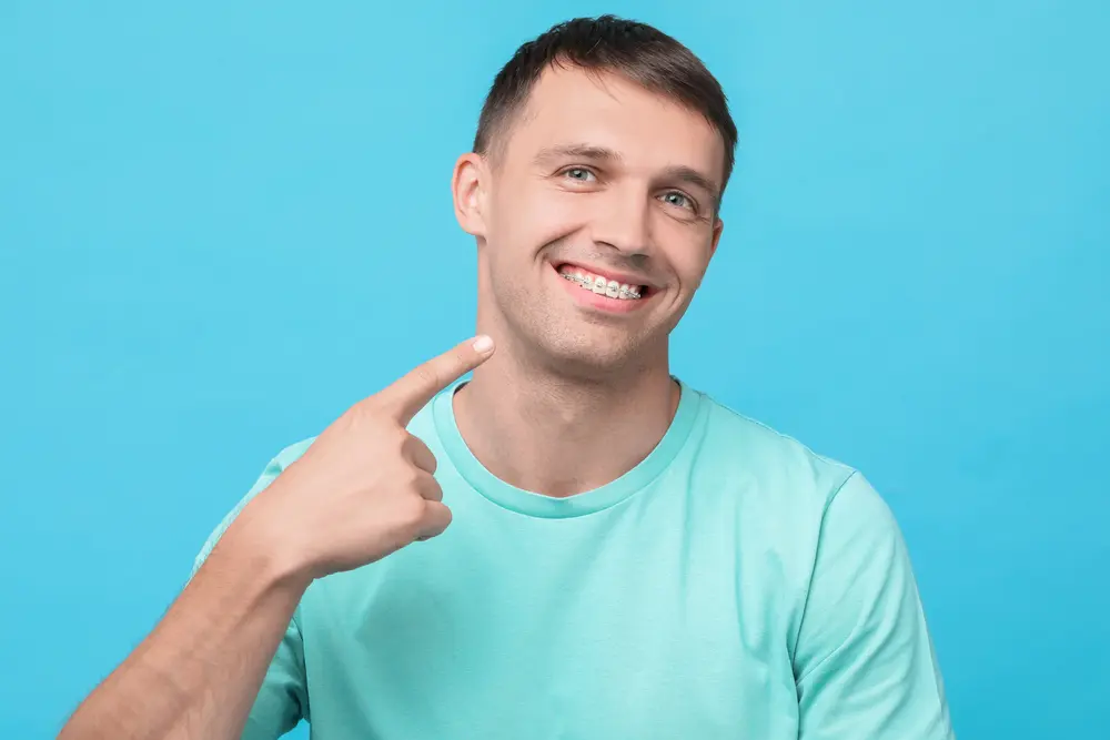 A man wearing a light blue shirt smiles and points at his metal braces, representing Levine Orthodontics in Fort Myers FL, against a solid blue background.