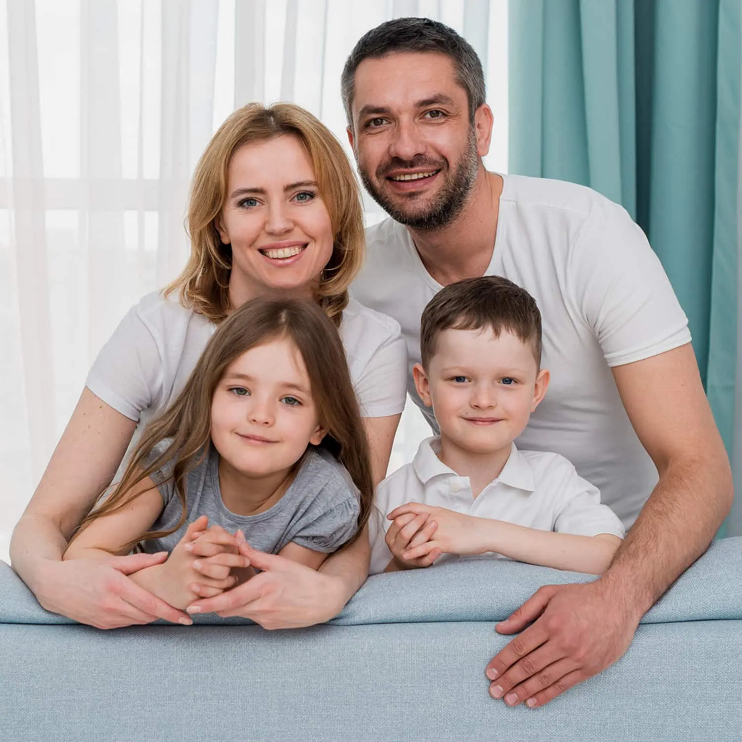 At Levine Orthodontics in Cape Coral, FL, a smiling family—man and woman seated closely behind a young girl and boy—hold hands on a light blue sofa, their orthodontic appliances adding sparkle to their joyful expressions in a bright room with white curtains and blue drapes.