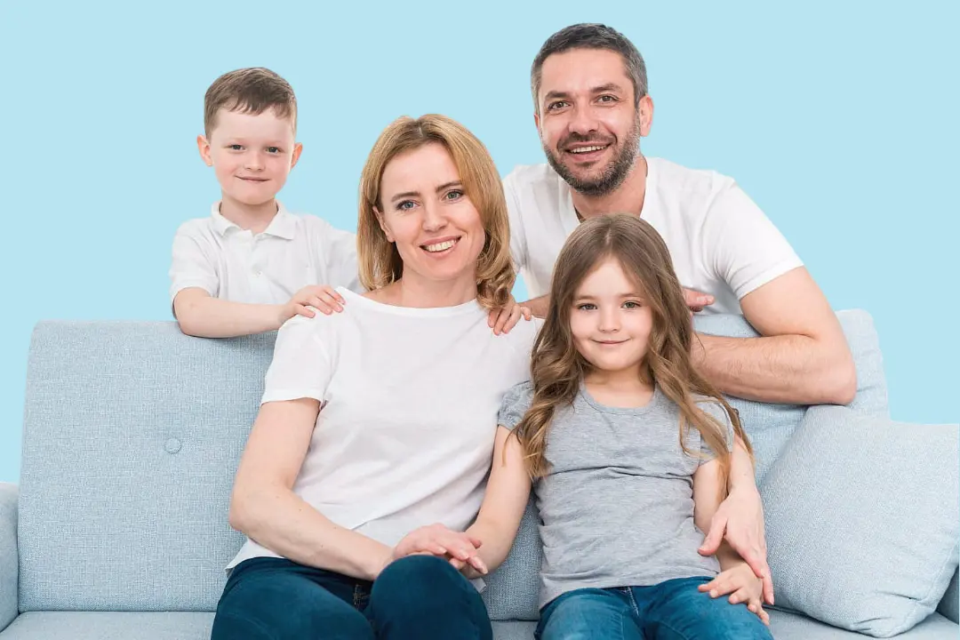 A smiling family of four, including one child displaying braces, sits together on a light blue couch against a blue background—capturing the care received at Levine Orthodontics in Fort Myers, Cape Coral, or Lehigh Acres, FL.