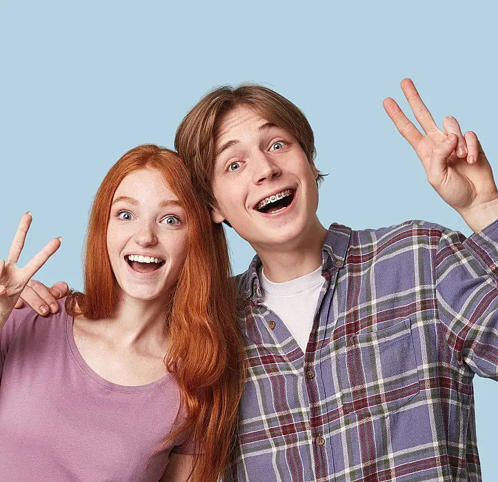 Two smiling teenagers with braces, patients of Levine Orthodontics in Fort Myers, FL, stand side by side against a light blue background, flashing peace signs.