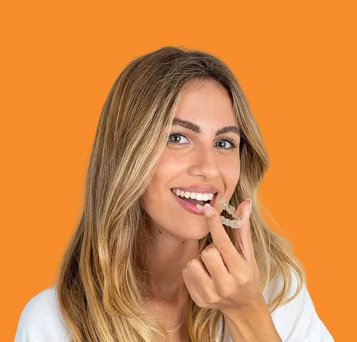 A woman with long blonde hair smiles and holds an Invisalign clear dental aligner near her mouth against an orange background, representing Levine Orthodontics in Fort Myers, FL.