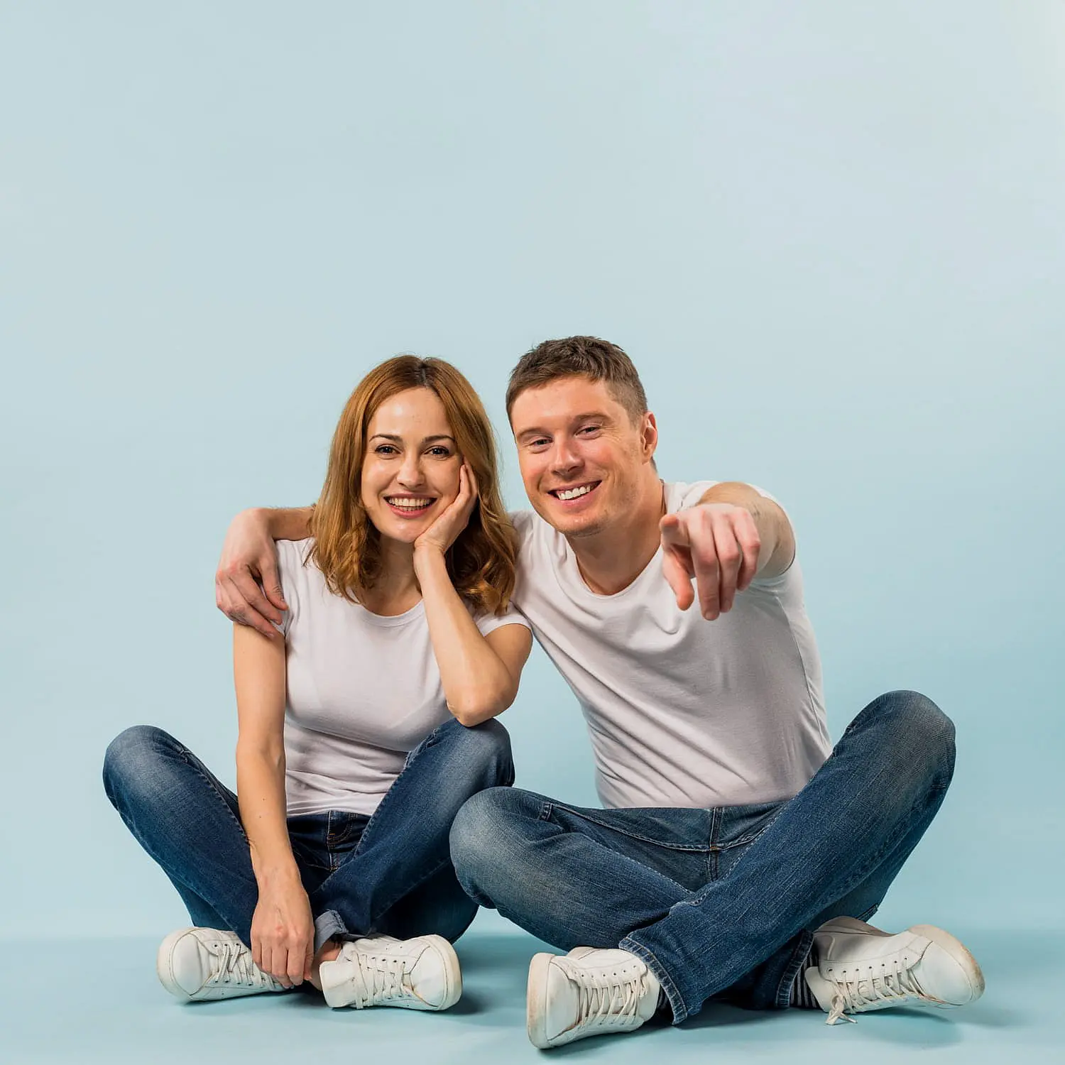 In a light blue setting, two people in white shirts and jeans sit together—one wears braces, smiling brightly with an arm around the other and pointing at the camera. This joyful moment captures the confidence that comes from treatment at Levine Orthodontics in Lehigh Acres, FL.