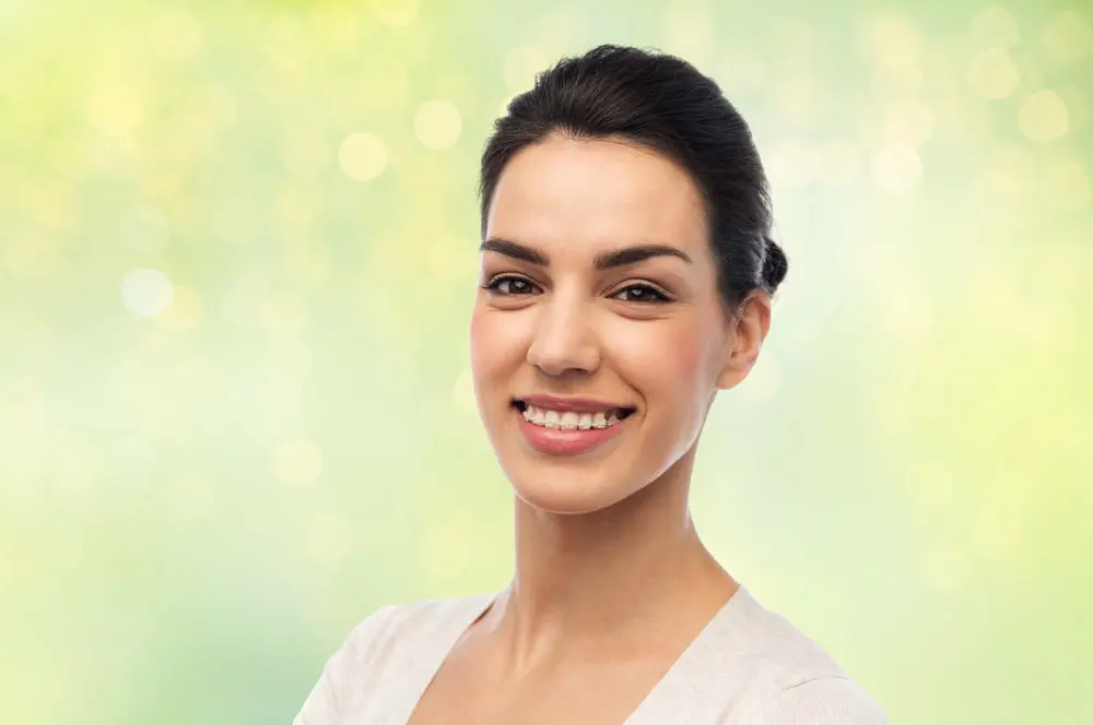 Smiling at the camera, a woman with dark hair tied back displays her clear braces in front of a soft, light green bokeh background—perfectly illustrating the confident results achievable with Levine Orthodontics in Lehigh Acres, FL.