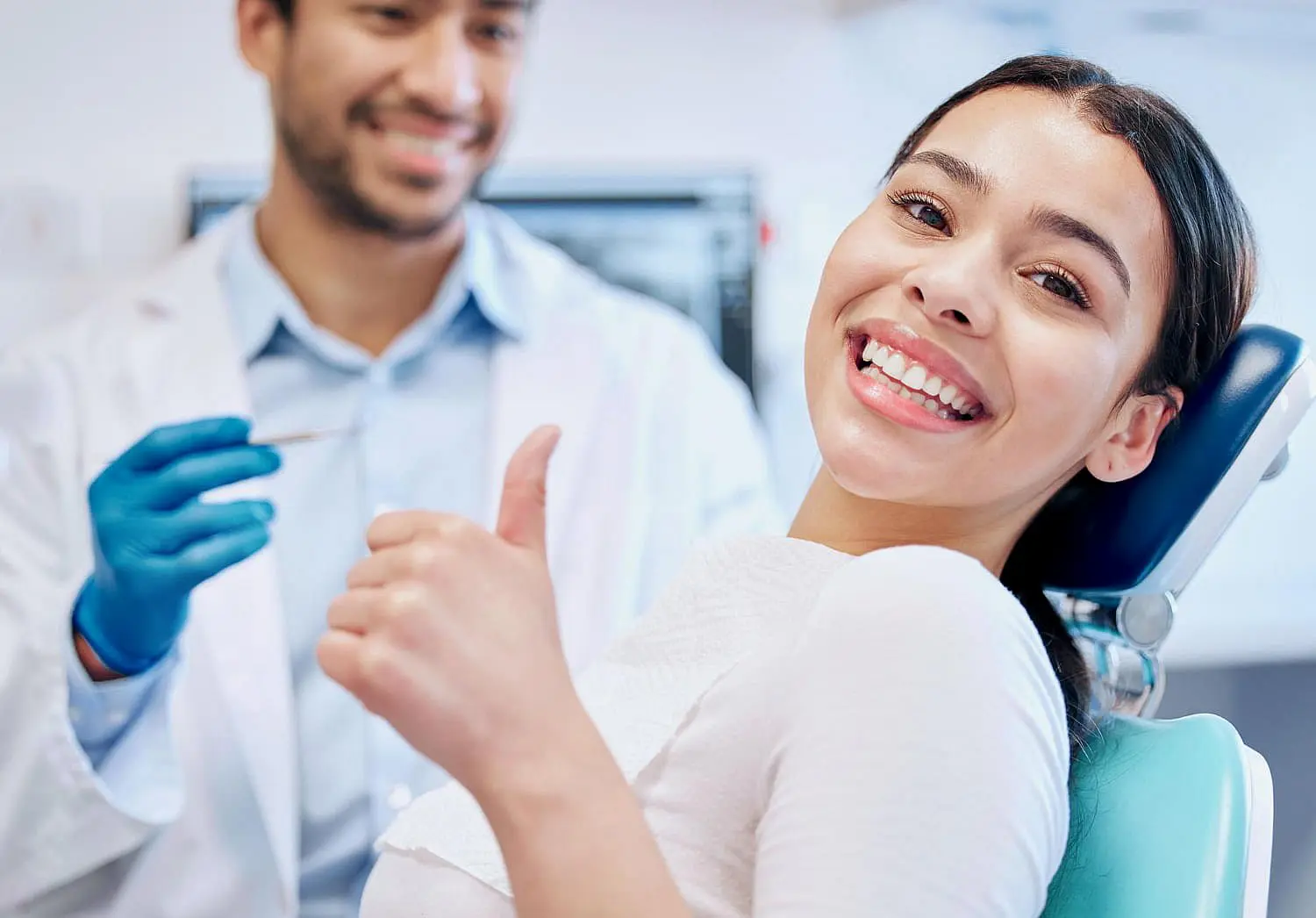 After completing Invisalign treatment at Levine Orthodontics in Lehigh Acres, FL, a woman confidently smiles and gives a thumbs-up from the dental chair as her dentist stands nearby with gloves and dental tools.
