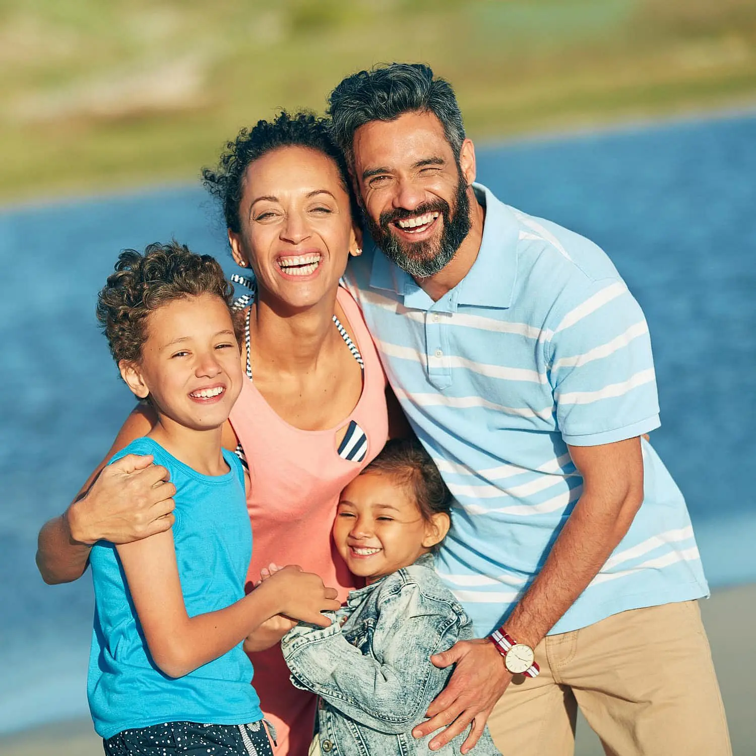A family of four with smiles outdoors near the water, with parents standing behind their two children—symbolizing adults and teens learning about Invisalign Clear Aligners, an ideal solution offered by Levine Orthodontics in Fort Myers, FL.