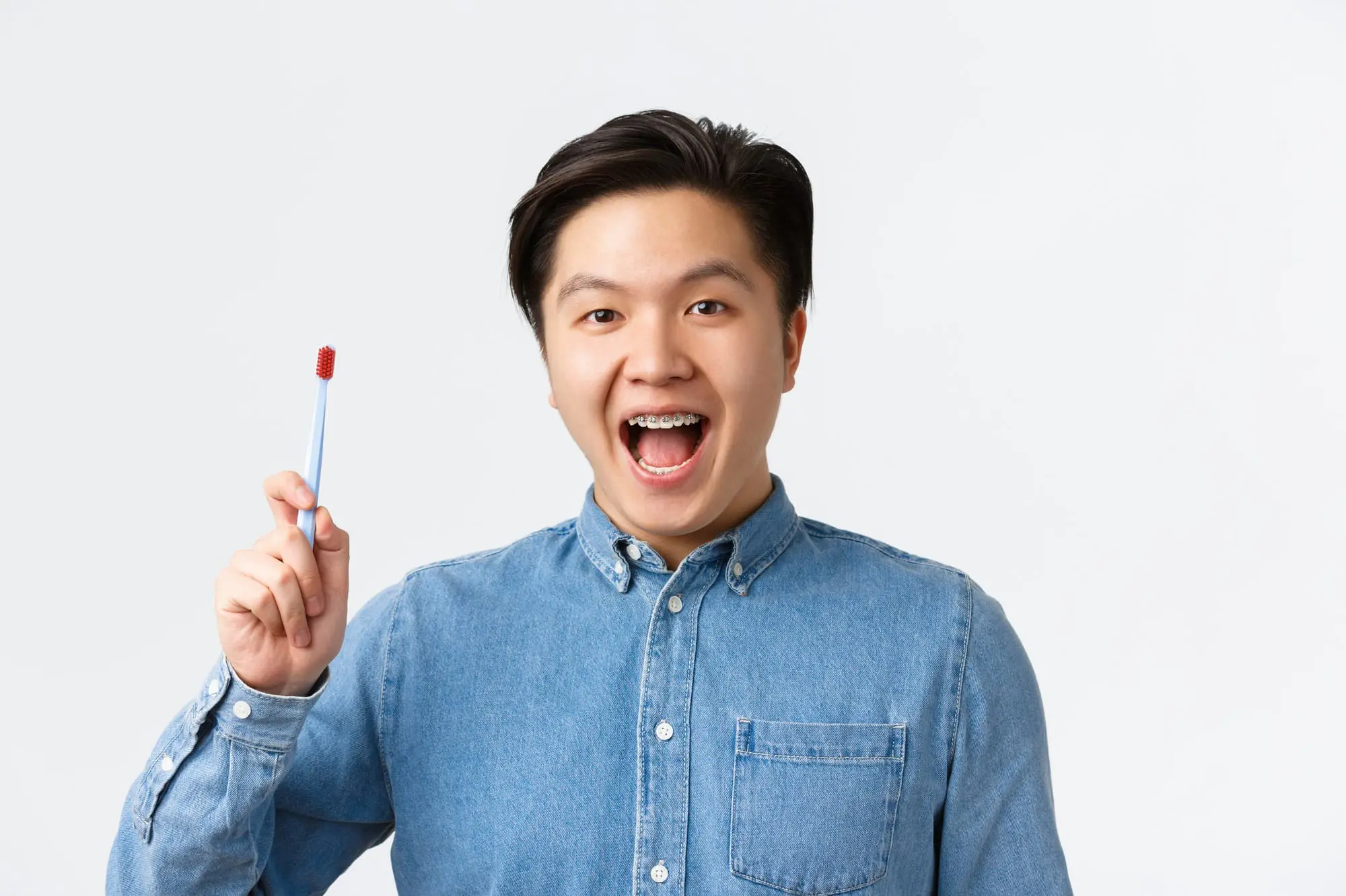 A person in a denim shirt, highlighting their braces while holding a toothbrush, represents adult orthodontic care with Levine Orthodontics in Fort Myers, Cape Coral or Lehigh Acres, FL, set against a simple light background.