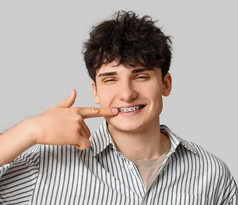 A young man in a striped shirt stands smiling against a plain gray background, pointing to his traditional metal braces received at Levine Orthodontics in Fort Myers, Cape Coral or Lehigh Acres, FL.