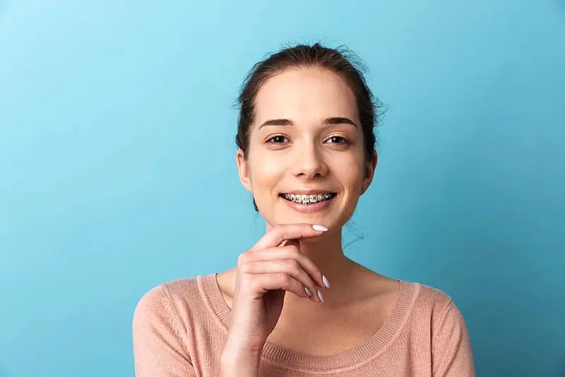 A young woman wearing metal braces and a light pink top smiles at the camera with her hand near her chin, pictured against a plain light blue background. This image represents patients at Levine Orthodontics in Lehigh Acres, FL.