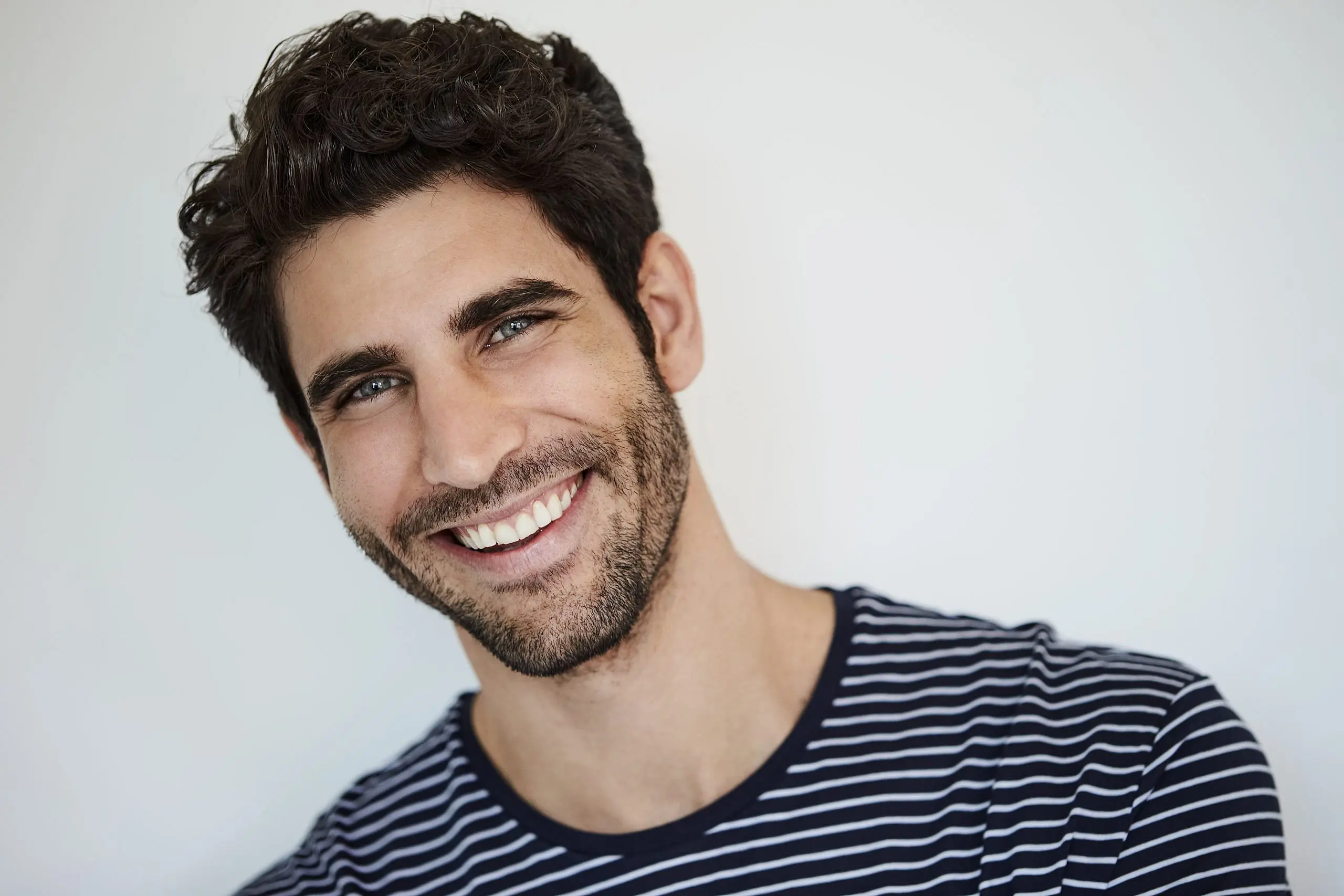 A man with dark hair and a beard, wearing a black and white striped shirt, smiles at the camera, revealing the subtle outline of a Palatal Expander in his teeth—an example of treatment available at Levine Orthodontics in Fort Myers, Cape Coral or Lehigh Acres, FL.