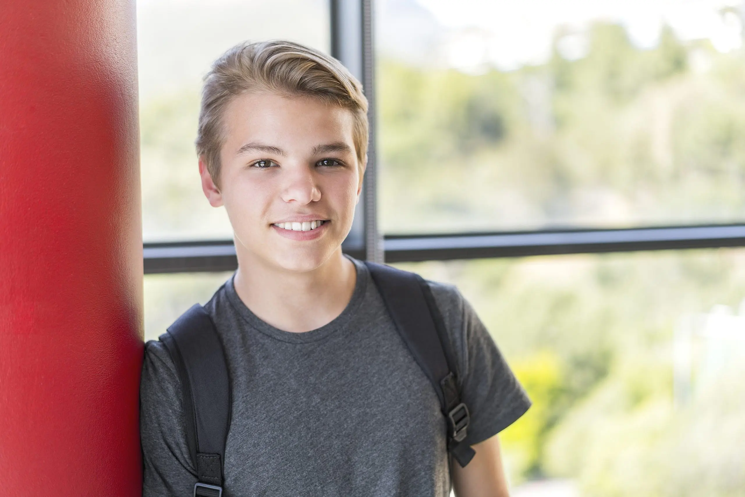 Smiling by a red column, a blonde teenage boy with braces confidently poses indoors in his gray t-shirt and backpack, large windows showcasing greenery behind him. Levine Orthodontics helps kids and teens achieve this confident smile in Fort Myers, Cape Coral or Lehigh Acres, FL—making every penny of braces worth it.