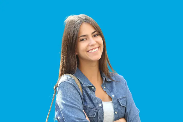 A young woman with long brown hair, wearing a denim jacket and a Herbst Appliance, smiles confidently with arms crossed against a solid blue background—showcasing the results of Levine Orthodontics in Fort Myers, Cape Coral, or Lehigh Acres, FL.