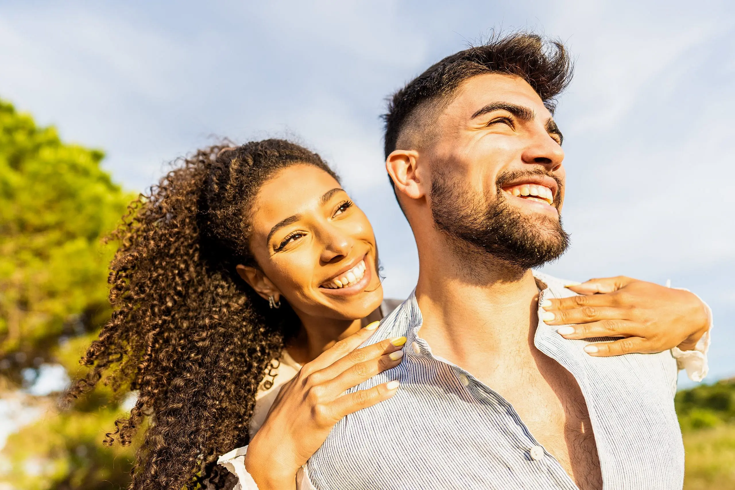 A happy couple enjoys a sunny day outdoors, surrounded by trees, with the woman’s clear braces from Levine Orthodontics in Fort Myers, Cape Coral, or Lehigh Acres, FL adding extra confidence to her radiant smile as she hugs her partner from behind.