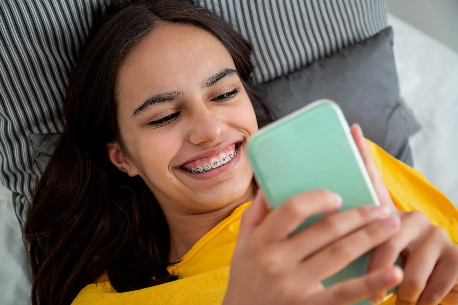 A teen girl with braces and a yellow shirt smiles at her smartphone while lying on a pillow, possibly exploring Braces Payment Plan options at Levine Orthodontics in Fort Myers, Cape Coral, or Lehigh Acres, FL.