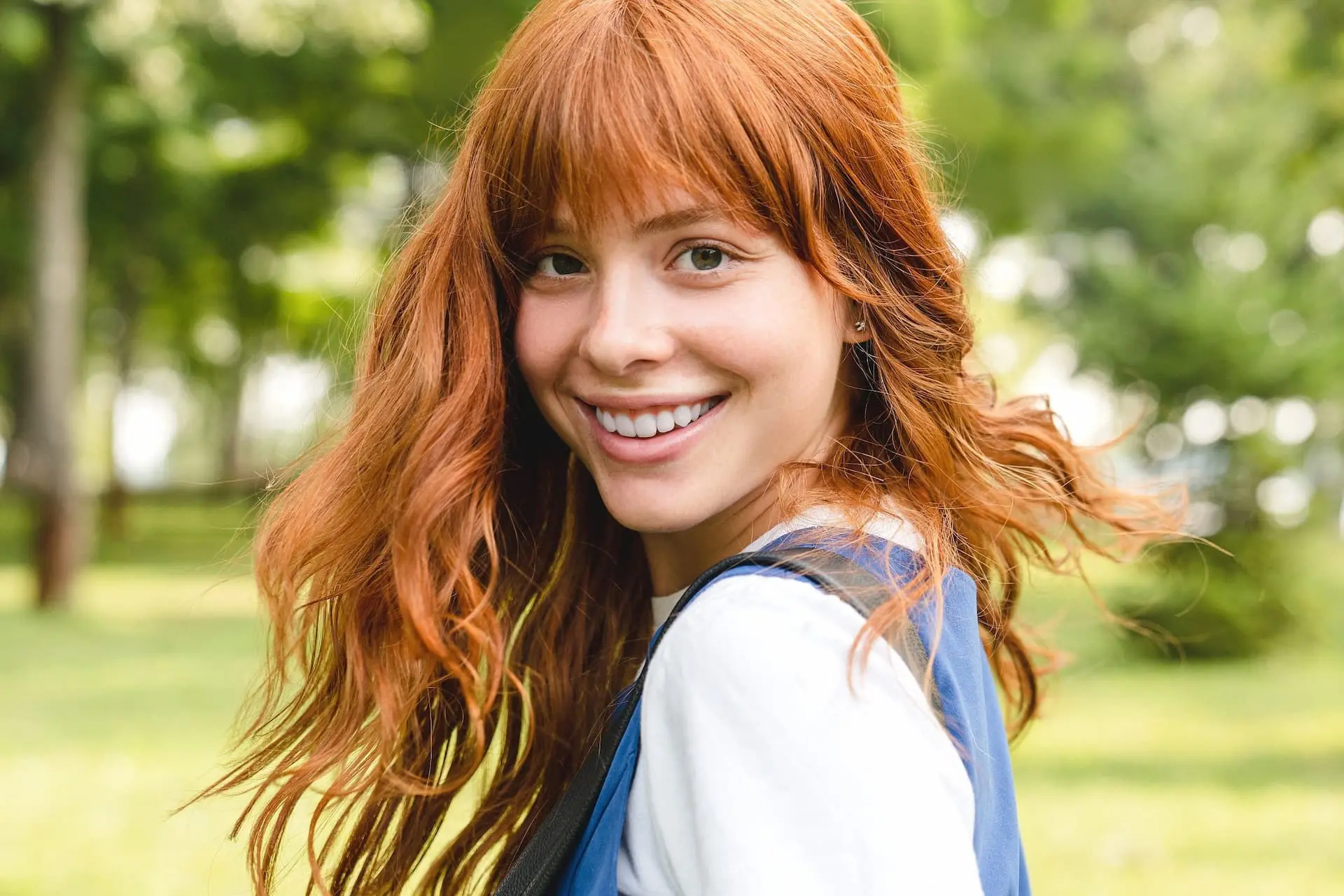 Smiling confidently in a green, leafy park, a person with long red hair and a blue top shows the assurance that comes from understanding Invisalign cost at Levine Orthodontics in Fort Myers, Cape Coral or Lehigh Acres, FL.