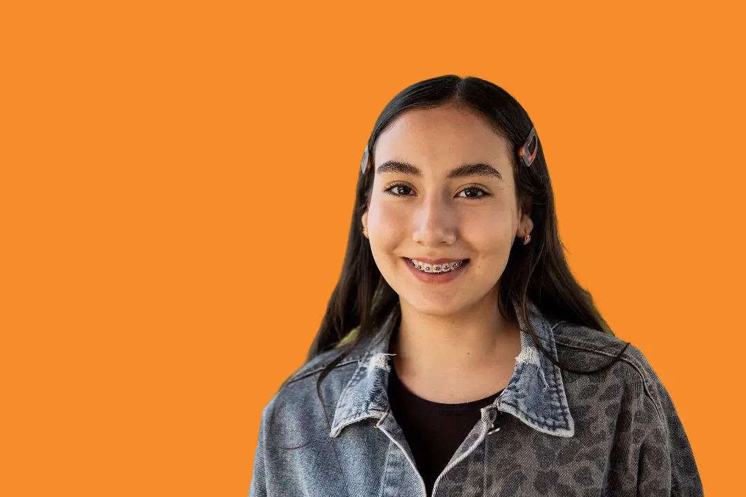 A teen girl with long dark hair, hair clips, a denim jacket, and braces flashes a smile against an orange background—representing Levine Orthodontics serving patients in Cape Coral, FL.