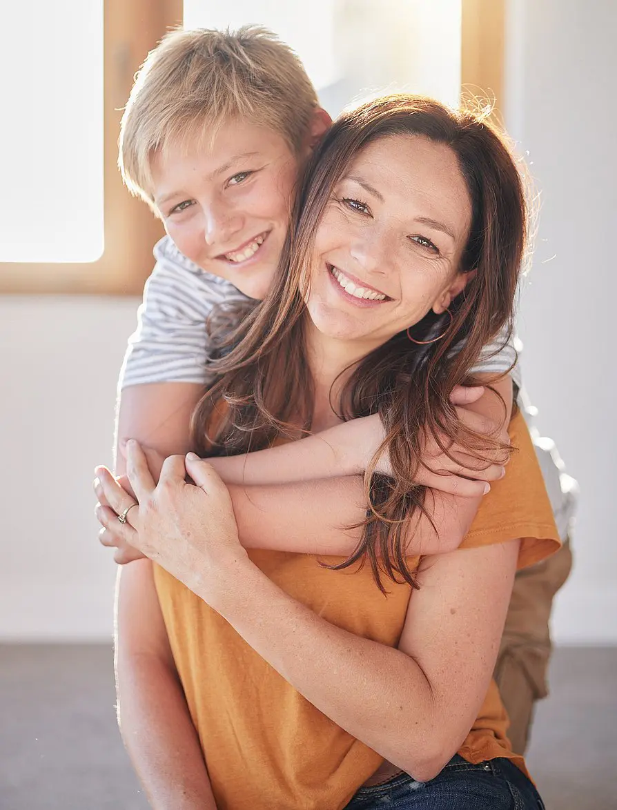 A young boy hugs a smiling woman from behind as they both face the camera in a brightly lit room, showcasing their confident smiles achieved with affordable Invisalign from Levine Orthodontics in Fort Myers, Cape Coral, or Lehigh Acres, FL.