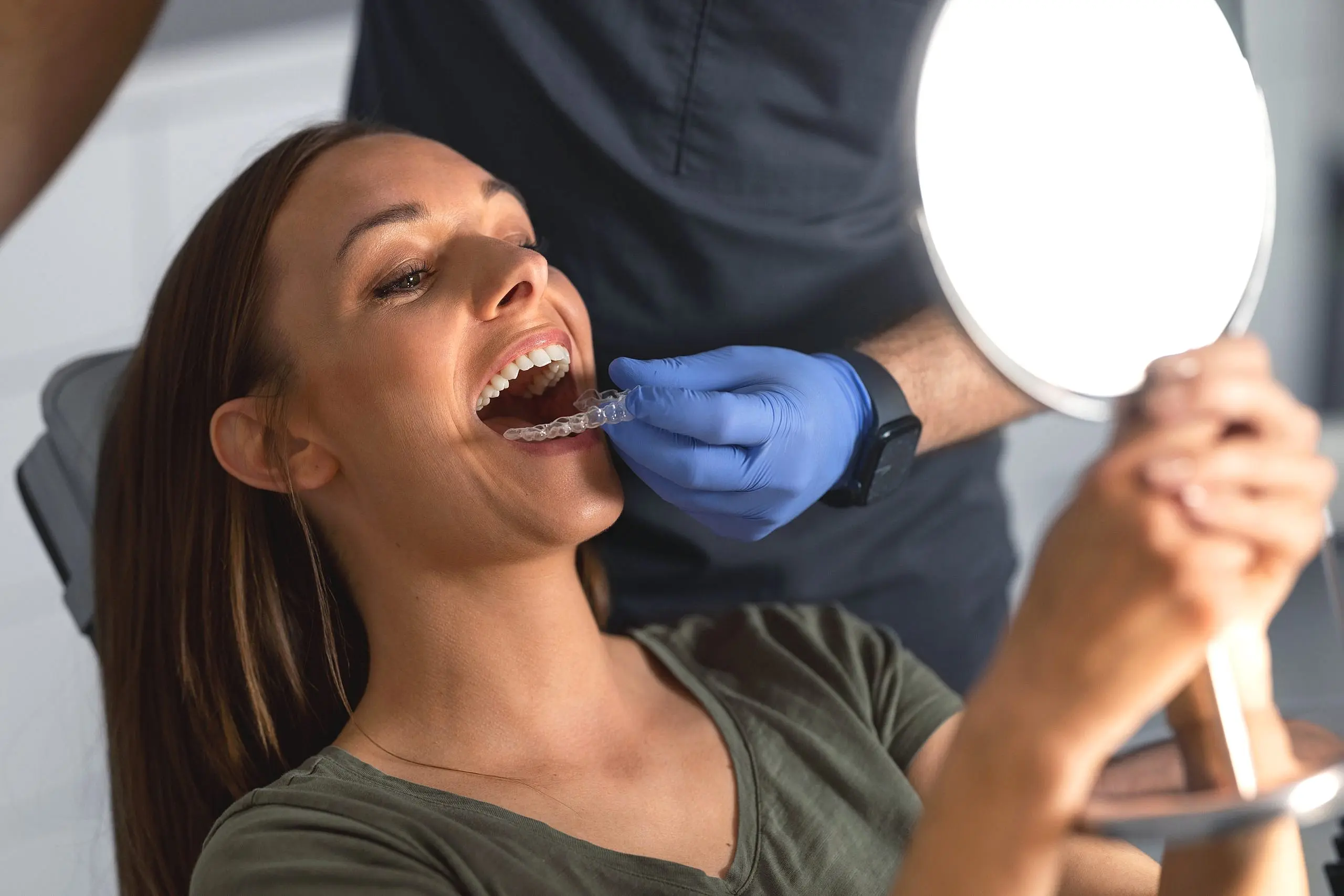 At Levine Orthodontics in Fort Myers, FL, a dentist fits a clear aligner onto a woman's teeth as she sits in the dental chair holding a mirror.