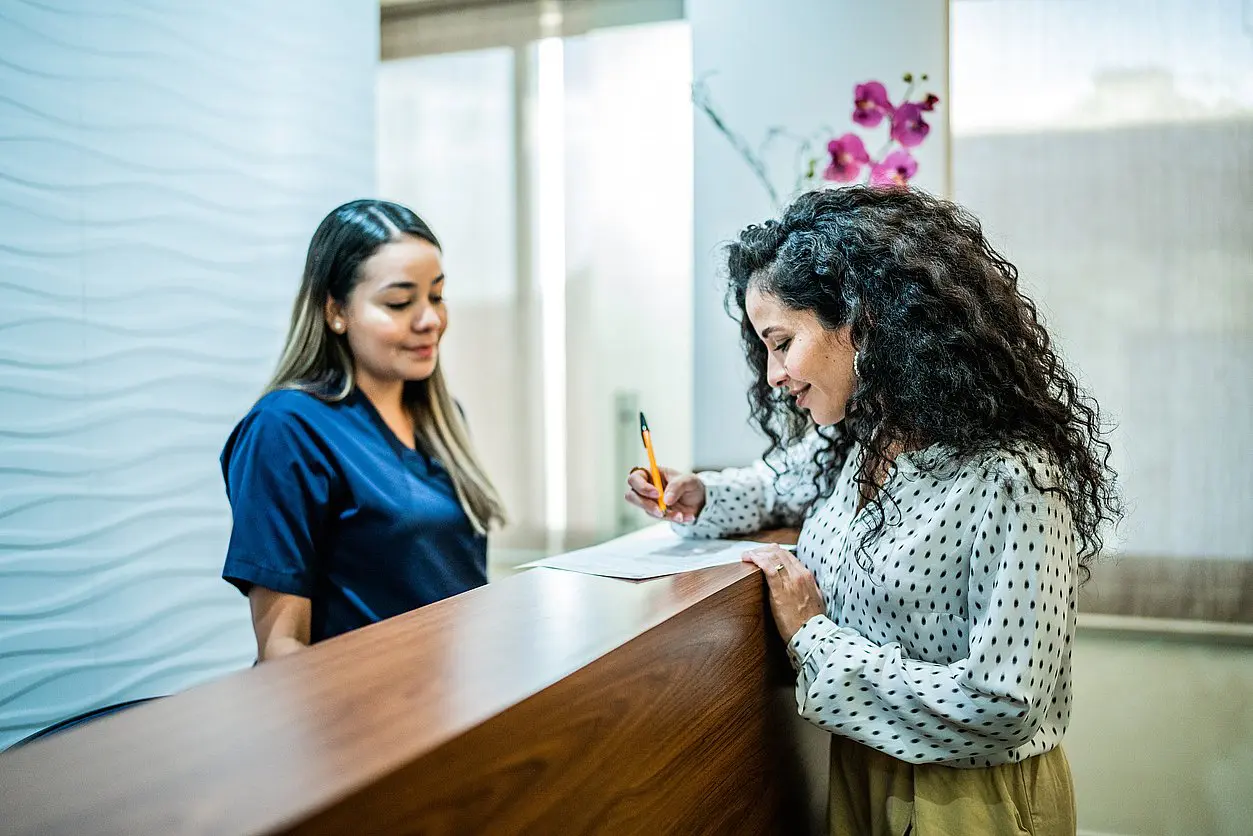 At Levine Orthodontics in Fort Myers, Cape Coral, or Lehigh Acres, FL, a woman completes paperwork at the reception desk as a receptionist in scrubs stands by to assist with Braces Payment Plan questions.