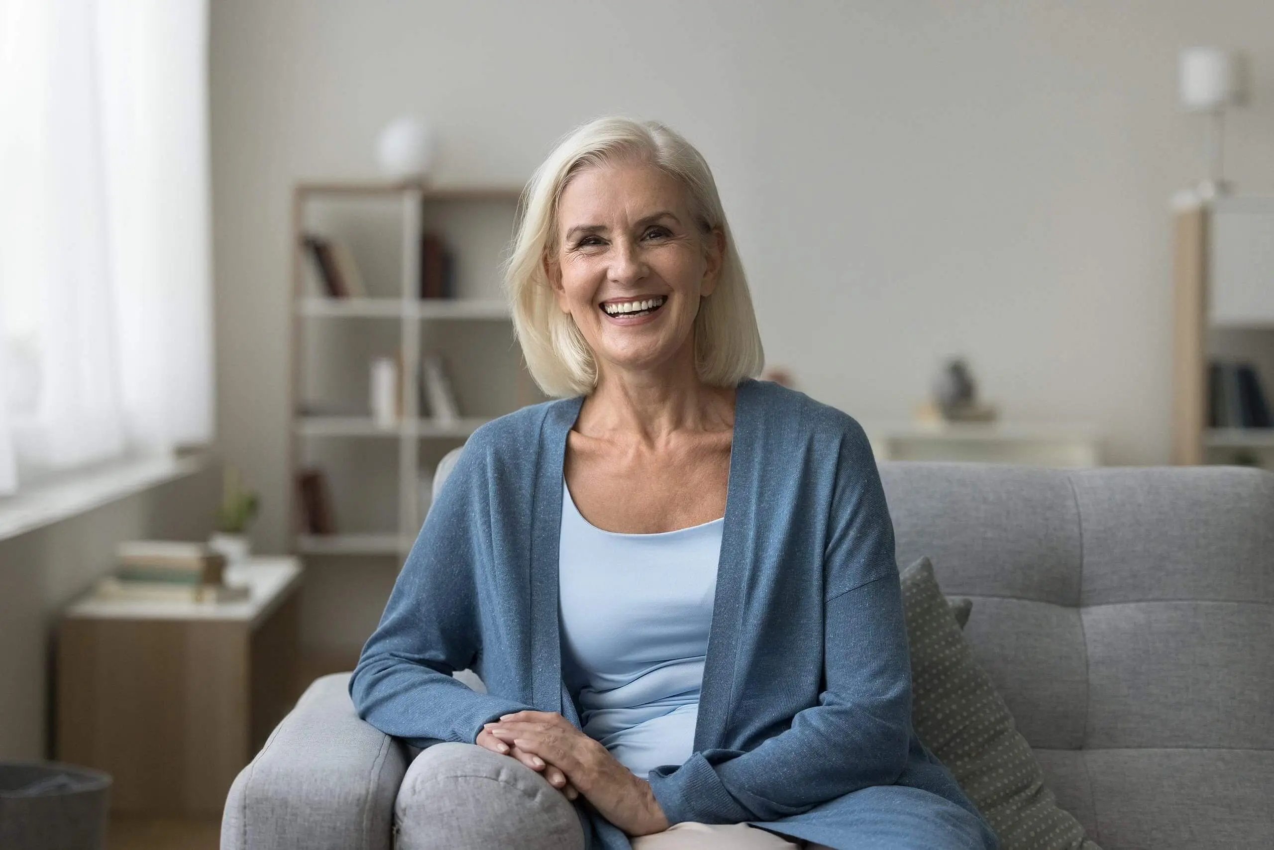 Considering Invisalign coverage under her dental plan, an older woman with gray hair, dressed in a light blue shirt and cardigan, sits smiling on a gray sofa in a bright living room with neatly lined shelves in the background. For those also exploring Invisalign options, Levine Orthodontics serves patients in Fort Myers, Cape Coral or Lehigh Acres, FL.
