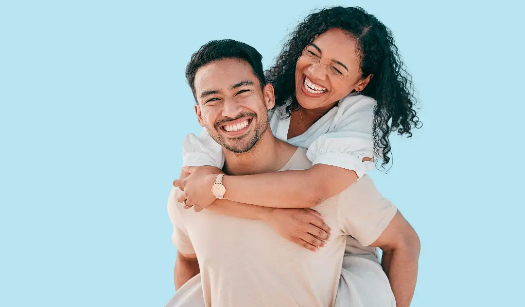 A man gives a piggyback ride to a smiling woman, both in light-colored outfits, as she confidently shows her Invisalign smile—courtesy of Levine Orthodontics in Lehigh Acres, FL—against a plain light blue background.