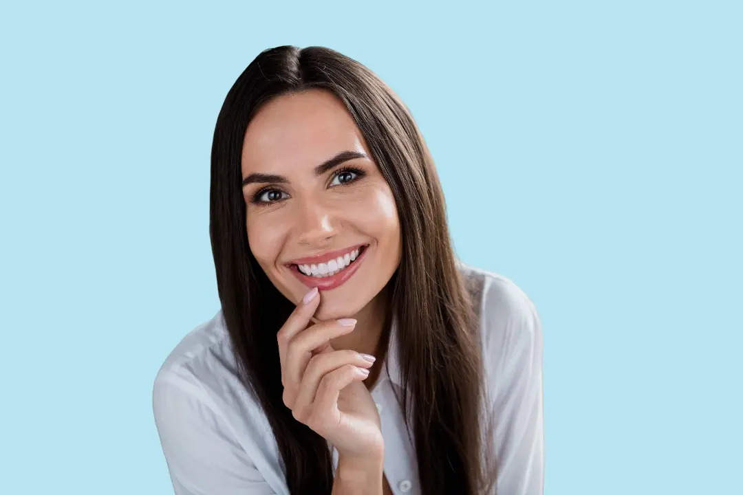 A woman with long brown hair in a white shirt smiles at the camera, resting her hand near her chin to show relief from jaw discomfort after using a TMJ Mouth Guard from Levine Orthodontics in Fort Myers, Cape Coral or Lehigh Acres, FL, set against a light blue background.