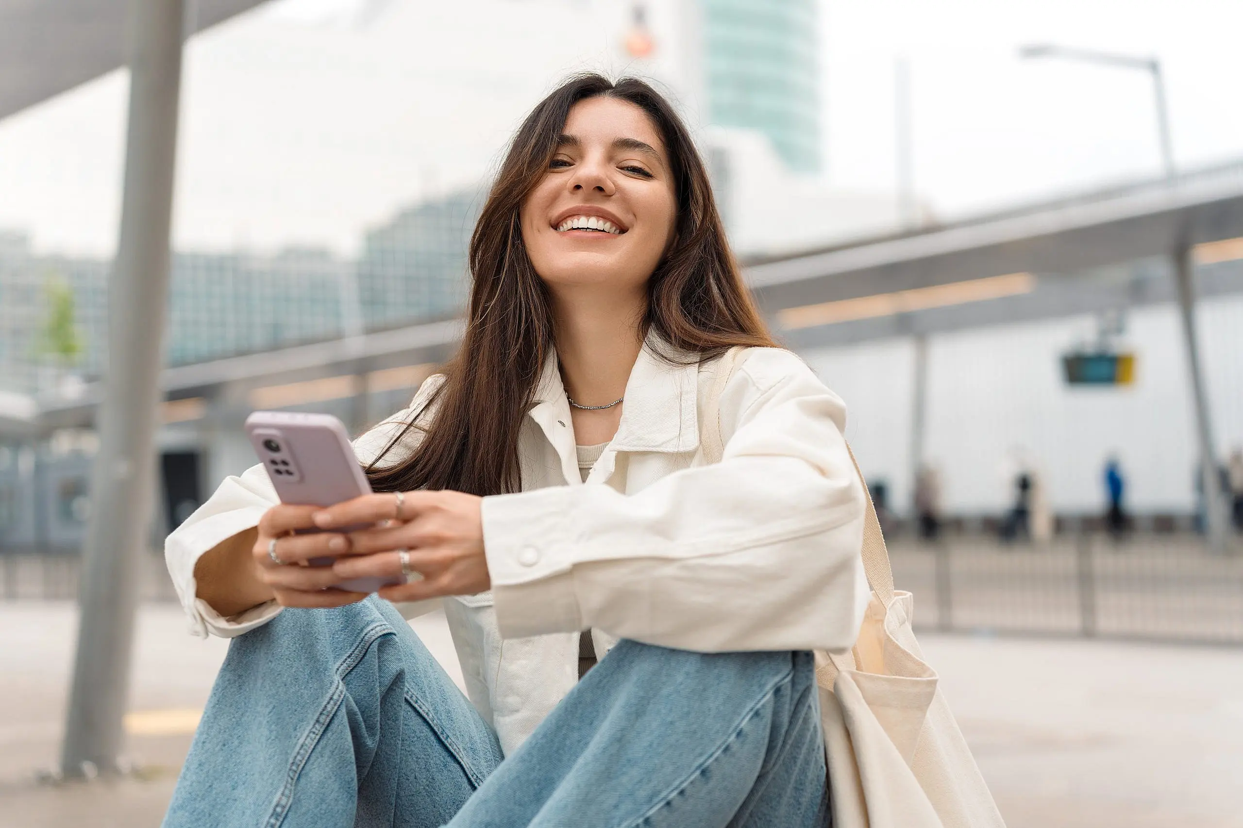 A young woman in a white jacket and jeans smiles while holding her smartphone outdoors against a blurred urban background, possibly exploring Invisalign Payment Plan options with Levine Orthodontics in Fort Myers, Cape Coral or Lehigh Acres, FL.