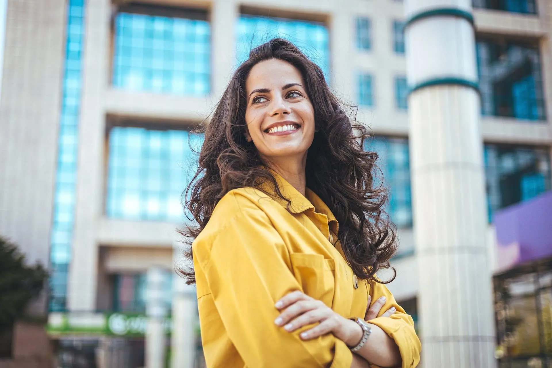 Confidently smiling with arms crossed, a woman in a yellow jacket stands outside a modern glass building, showcasing her bright smile after discovering the affordable braces cost at Levine Orthodontics in Fort Myers, Cape Coral or Lehigh Acres, FL.