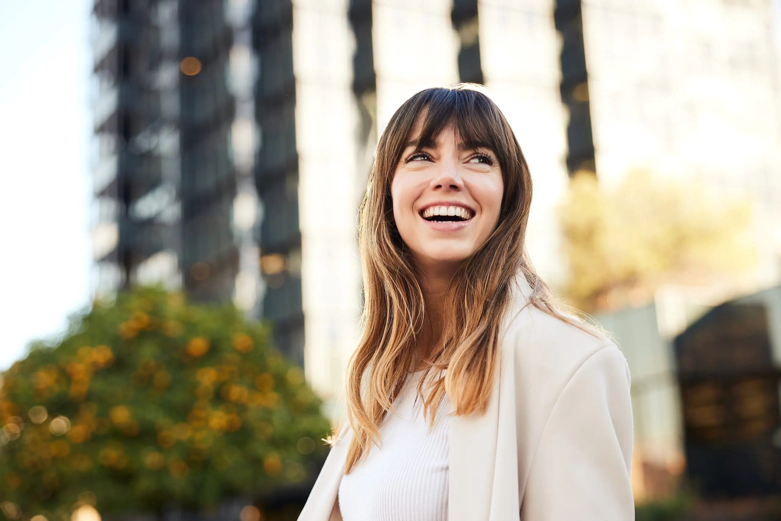 Smiling confidently with affordable braces, a woman with long brown hair and bangs stands outdoors near a modern glass building and trees—showcasing the work of Levine Orthodontics in Fort Myers, Cape Coral or Lehigh Acres, FL.