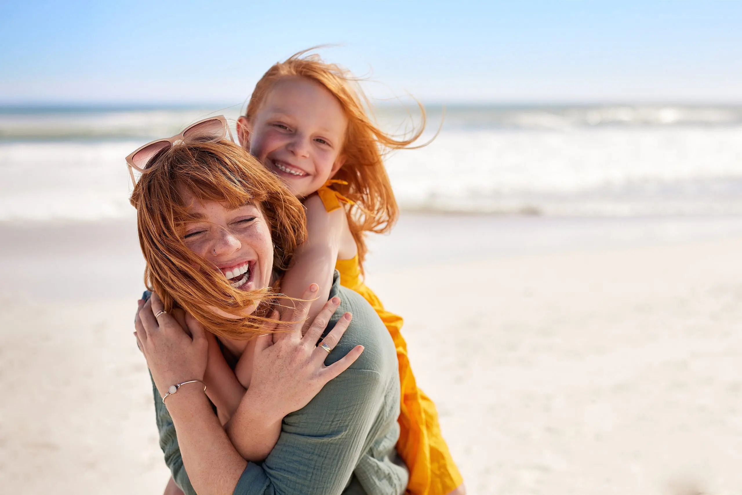 A woman gives a smiling young girl with metal braces a piggyback ride on a sandy beach, their hair windswept and ocean waves in the background—all moments you can enjoy after visiting Levine Orthodontics in Fort Myers, Cape Coral, or Lehigh Acres, FL.