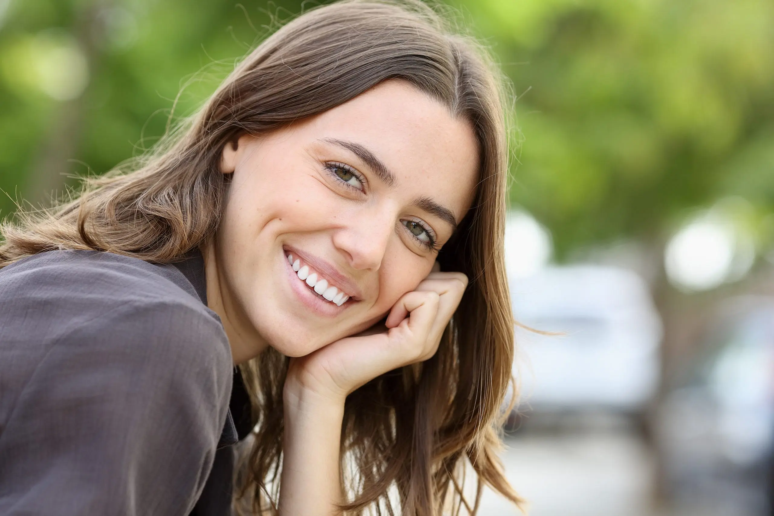 Smiling confidently at the camera, a woman with long brown hair in a gray shirt displays her affordable braces against a backdrop of blurred green trees—an example of the beautiful results possible at Levine Orthodontics in Fort Myers, Cape Coral or Lehigh Acres, FL.