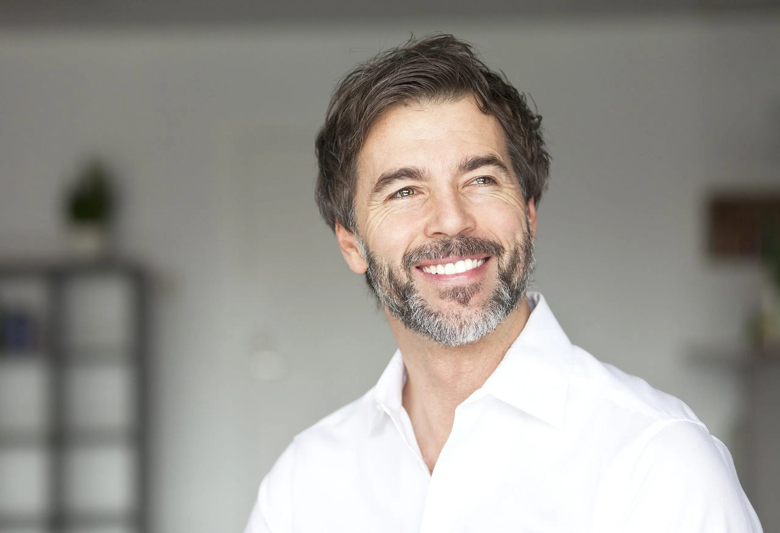 A man with short brown hair and a beard, wearing a white shirt, smiles and shows off his clear braces in a bright indoor setting—an example of the confident results possible at Levine Orthodontics in Fort Myers, Cape Coral or Lehigh Acres, FL.