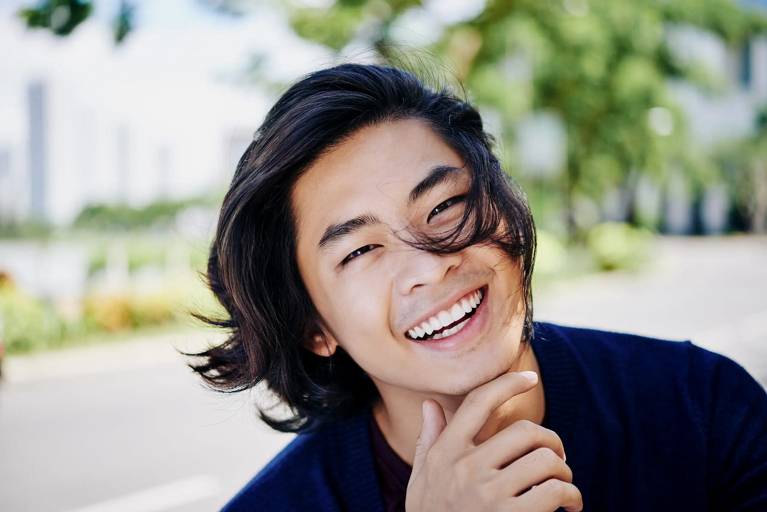 Confident in their smile thanks to affordable braces from Levine Orthodontics in Fort Myers, Cape Coral, or Lehigh Acres, FL, a young person with long dark hair smiles outdoors, resting their chin on one hand against a backdrop of blurred greenery and buildings.