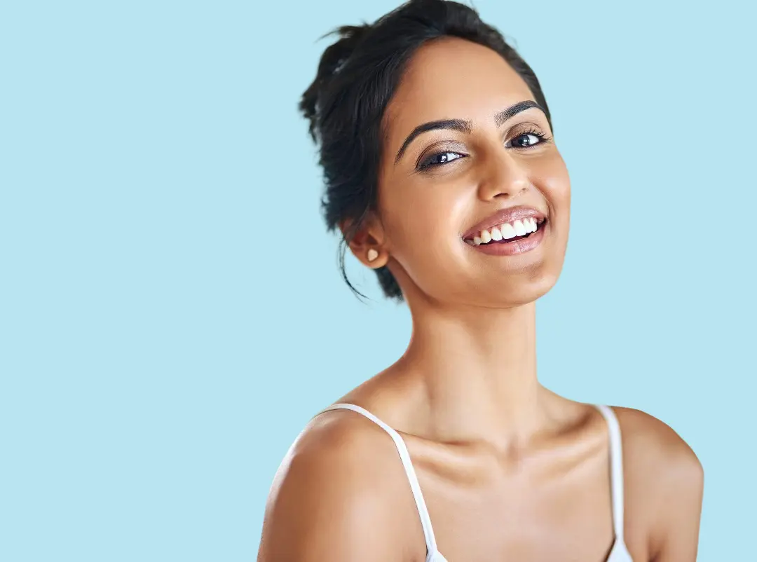 Smiling confidently in a white tank top, a dark-haired woman displays her gold braces for Levine Orthodontics in Fort Myers, Cape Coral, or Lehigh Acres, FL, set against a light blue background.