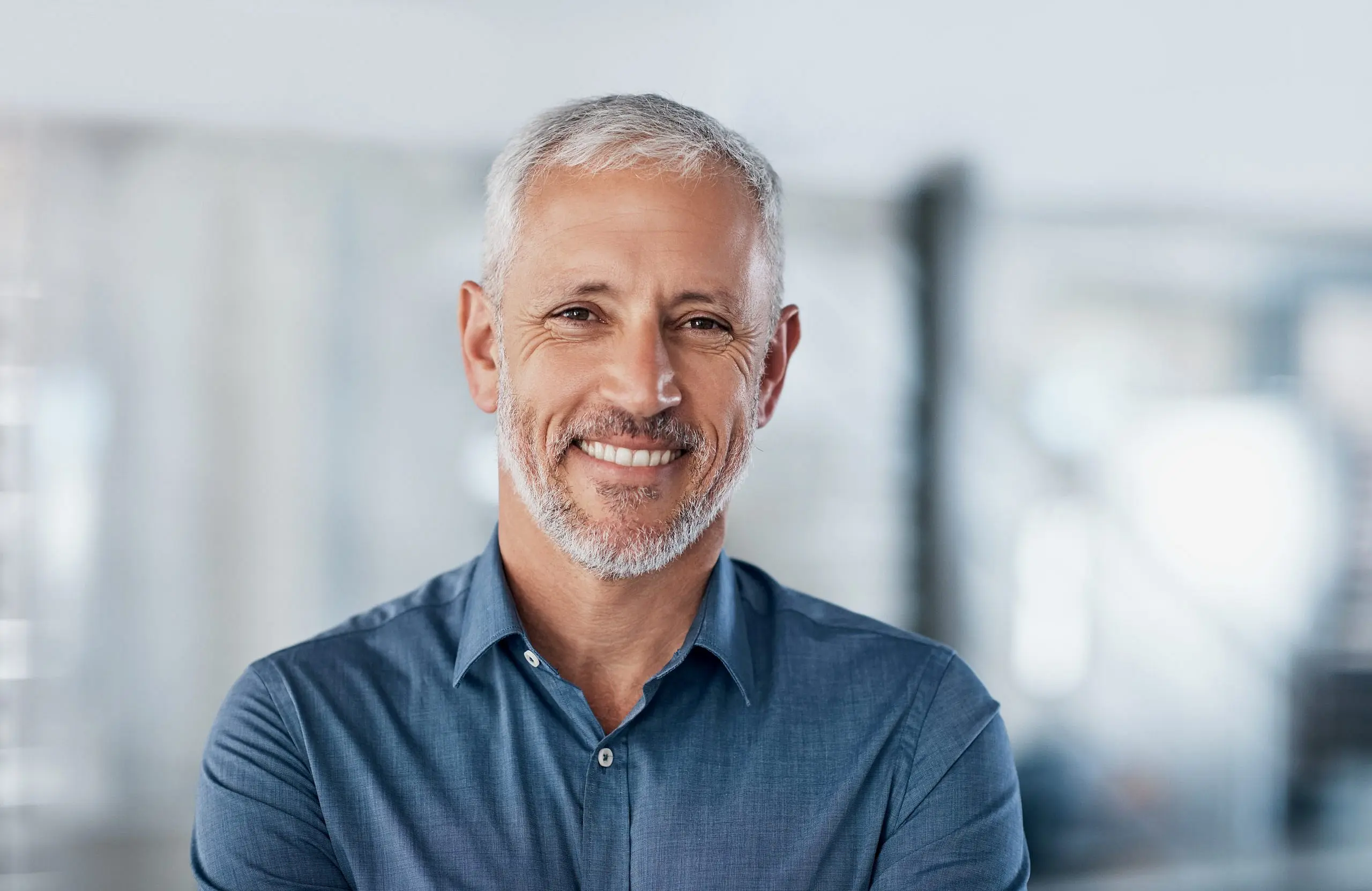 Feeling confident after discovering affordable braces cost options at Levine Orthodontics in Fort Myers, Cape Coral, or Lehigh Acres, FL, a middle-aged man with short gray hair and beard smiles with arms crossed in a brightly lit indoor space.