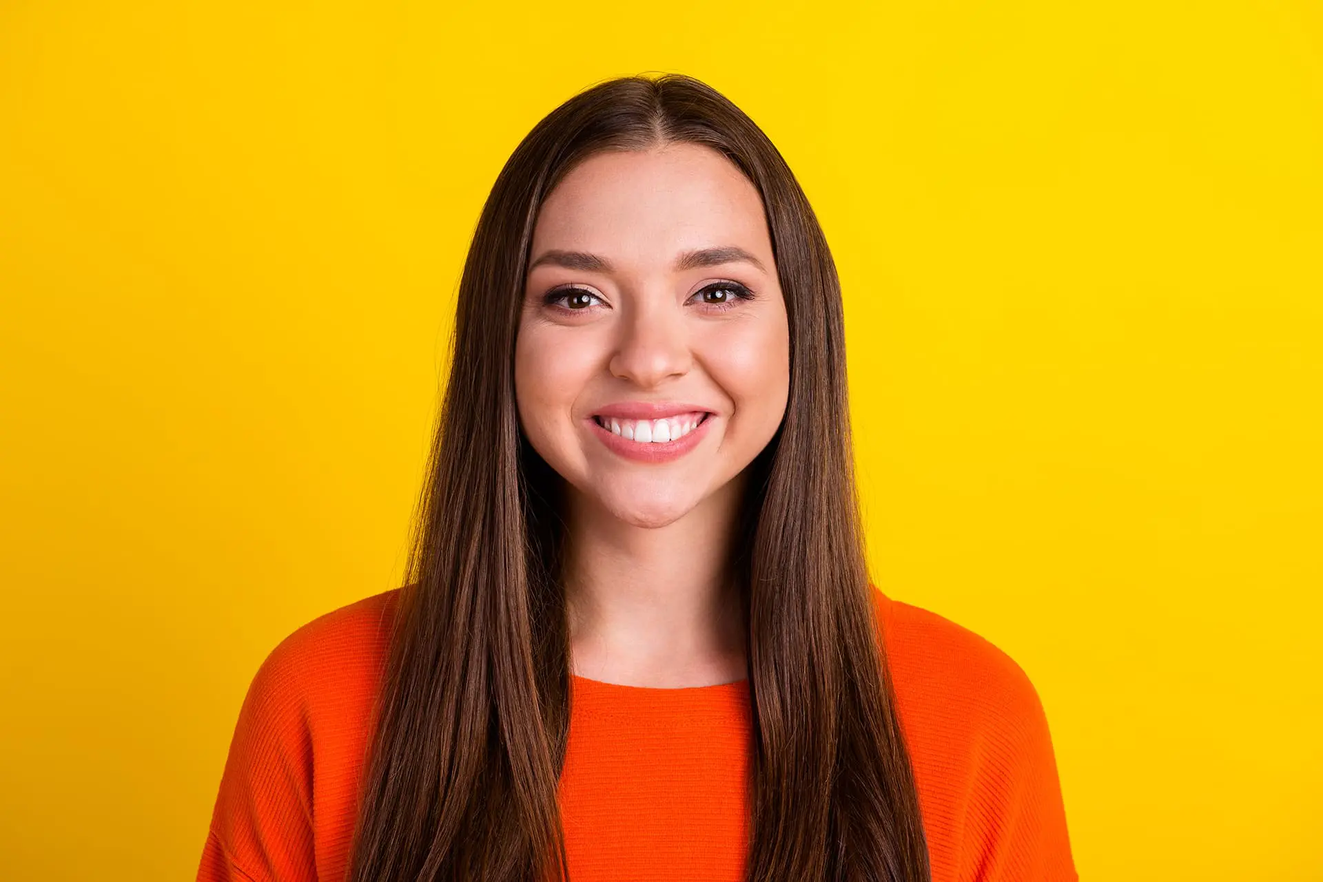 A young woman with long brown hair, wearing an orange top, smiles confidently—possibly due to different types of braces from Levine Orthodontics in Fort Myers, Cape Coral or Lehigh Acres, FL—against a solid yellow background.