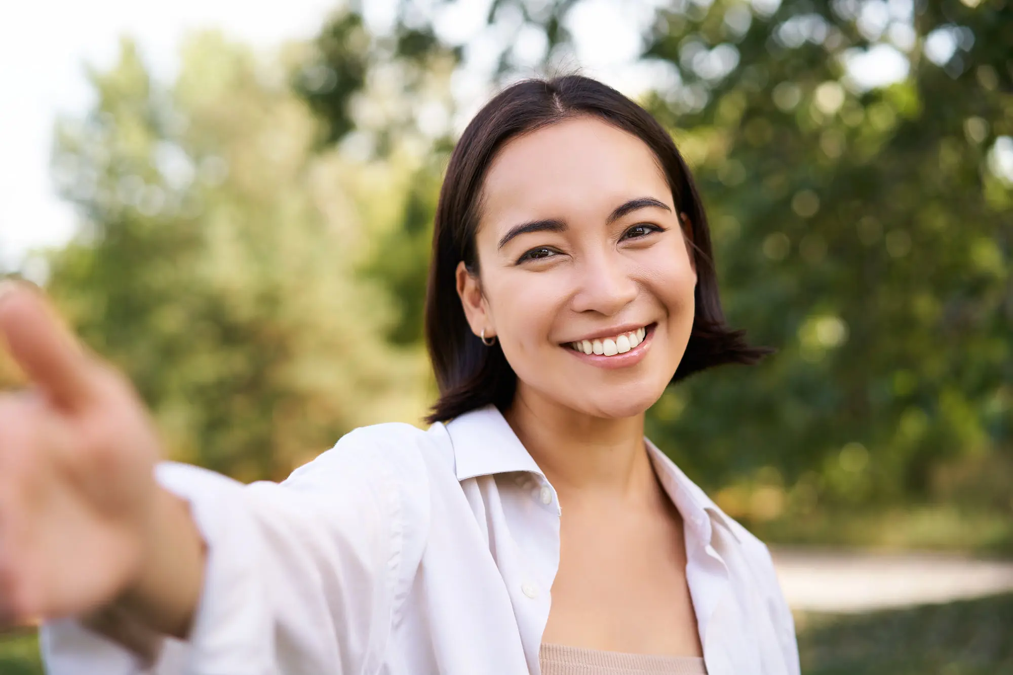 Displaying the confidence that comes from using orthodontic appliances at Levine Orthodontics in Fort Myers, Cape Coral or Lehigh Acres, FL, a smiling woman outdoors reaches toward the camera with greenery and trees softly blurred behind her.