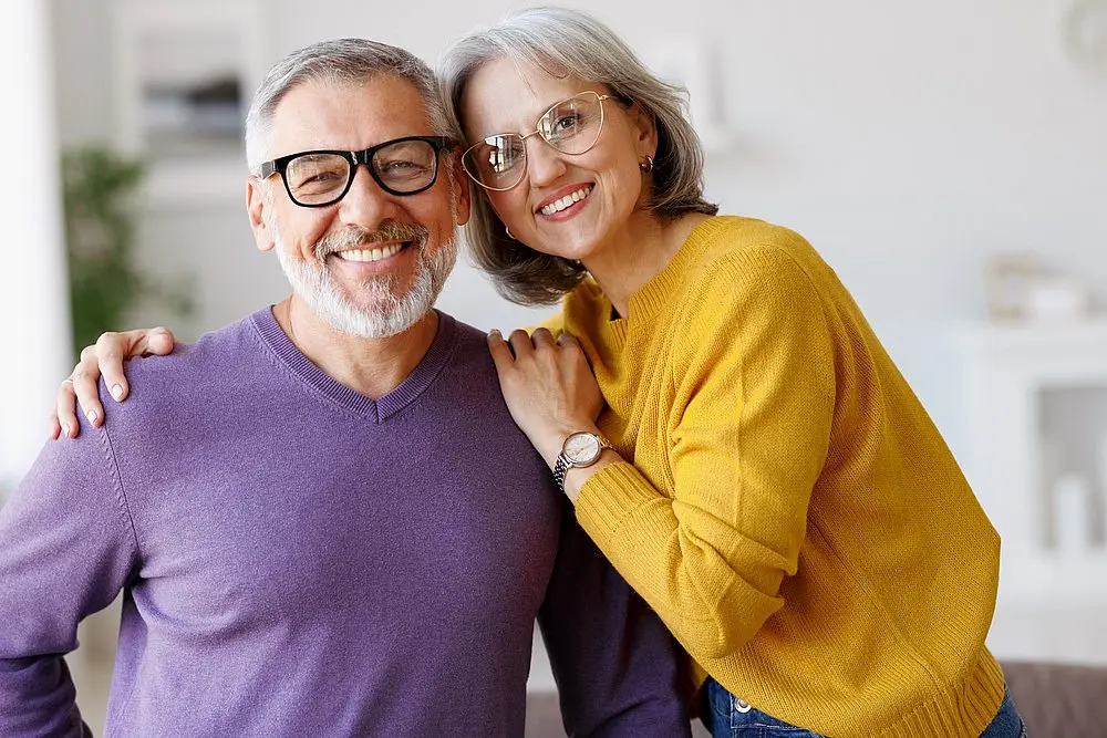 At Levine Orthodontics in Fort Myers, Cape Coral, or Lehigh Acres, FL, an older man in a purple sweater and glasses stands smiling next to an older woman in a yellow sweater and glasses indoors.