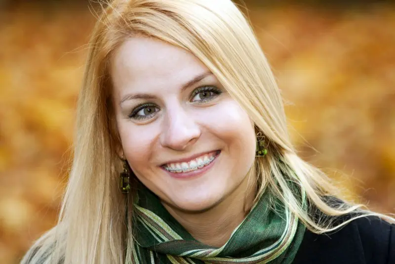 A woman with long blond hair, wearing a green scarf and earrings, shows off her clear ceramic braces in an autumn setting—an example of the confident smiles created by Levine Orthodontics in Lehigh Acres, FL.