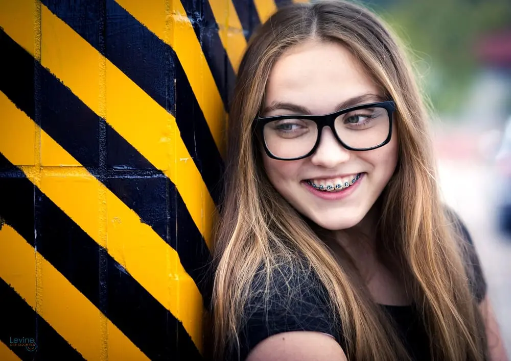A teen girl with long brown hair and glasses smiles with braces from Levine Orthodontics correcting her overbite, standing by a yellow and black striped wall in Fort Myers, Cape Coral or Lehigh Acres, FL.