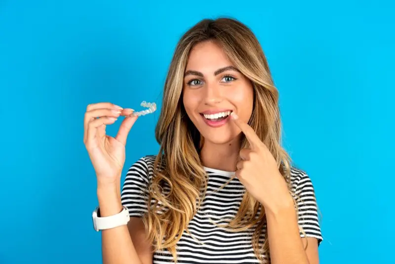 Highlighting the benefits of Orthodontic Insurance Plans, a woman in a striped shirt smiles and points at her teeth while holding a clear dental aligner—all services available at Levine Orthodontics in Fort Myers, Cape Coral or Lehigh Acres, FL.