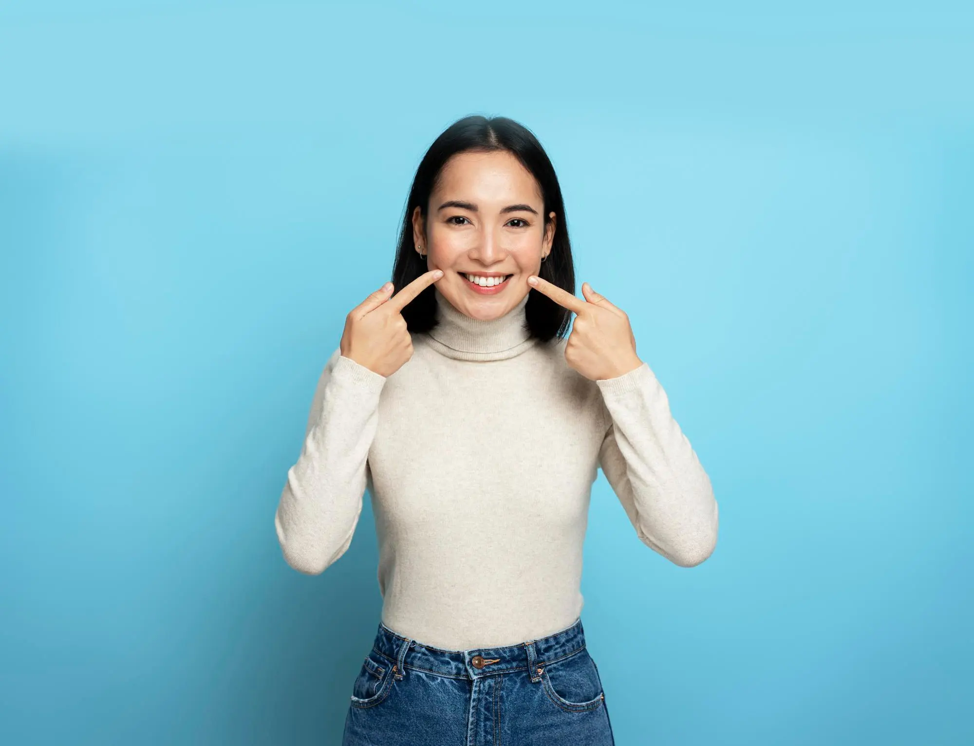 Smiling and pointing at her mouth, a woman in a beige turtleneck and jeans poses against a blue background, representing Levine Orthodontics in Fort Myers, Cape Coral or Lehigh Acres, FL.