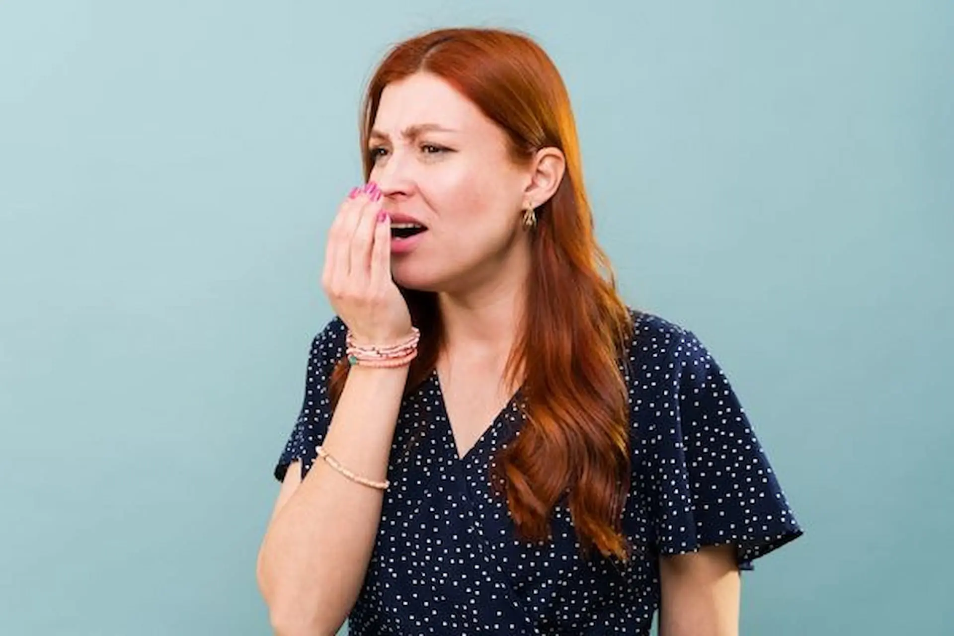 A woman with long red hair in a navy shirt appears uncomfortable, covering her mouth with her hand against a plain teal background—an image representative of the concern some patients feel before visiting Levine Orthodontics in Fort Myers, Cape Coral, or Lehigh Acres, FL.