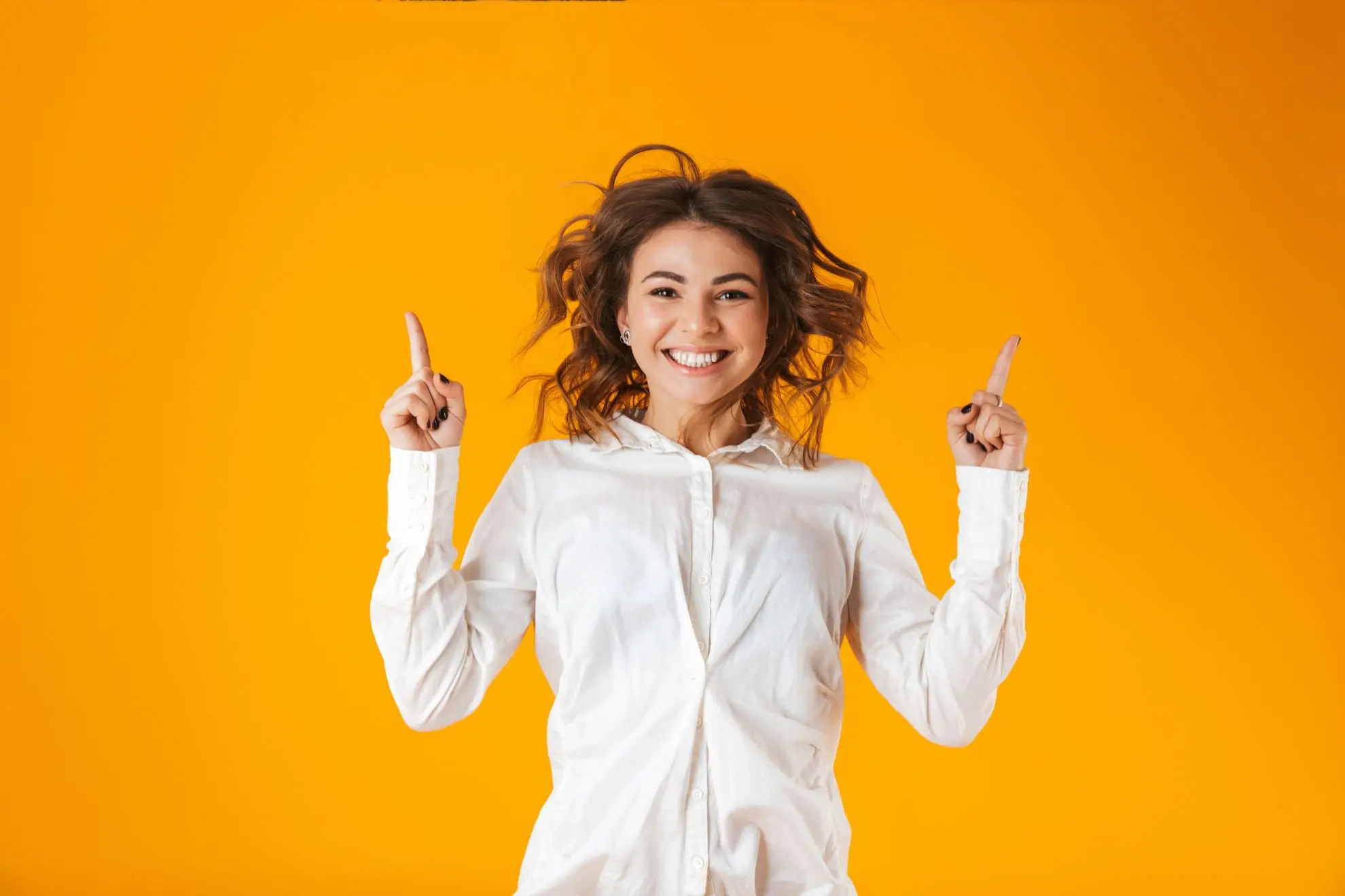 A young woman in a white shirt smiles and points upward with both index fingers against a solid orange background, proudly showing her Invisalign results with Levine Orthodontics in Fort Myers, Cape Coral, or Lehigh Acres, FL.
