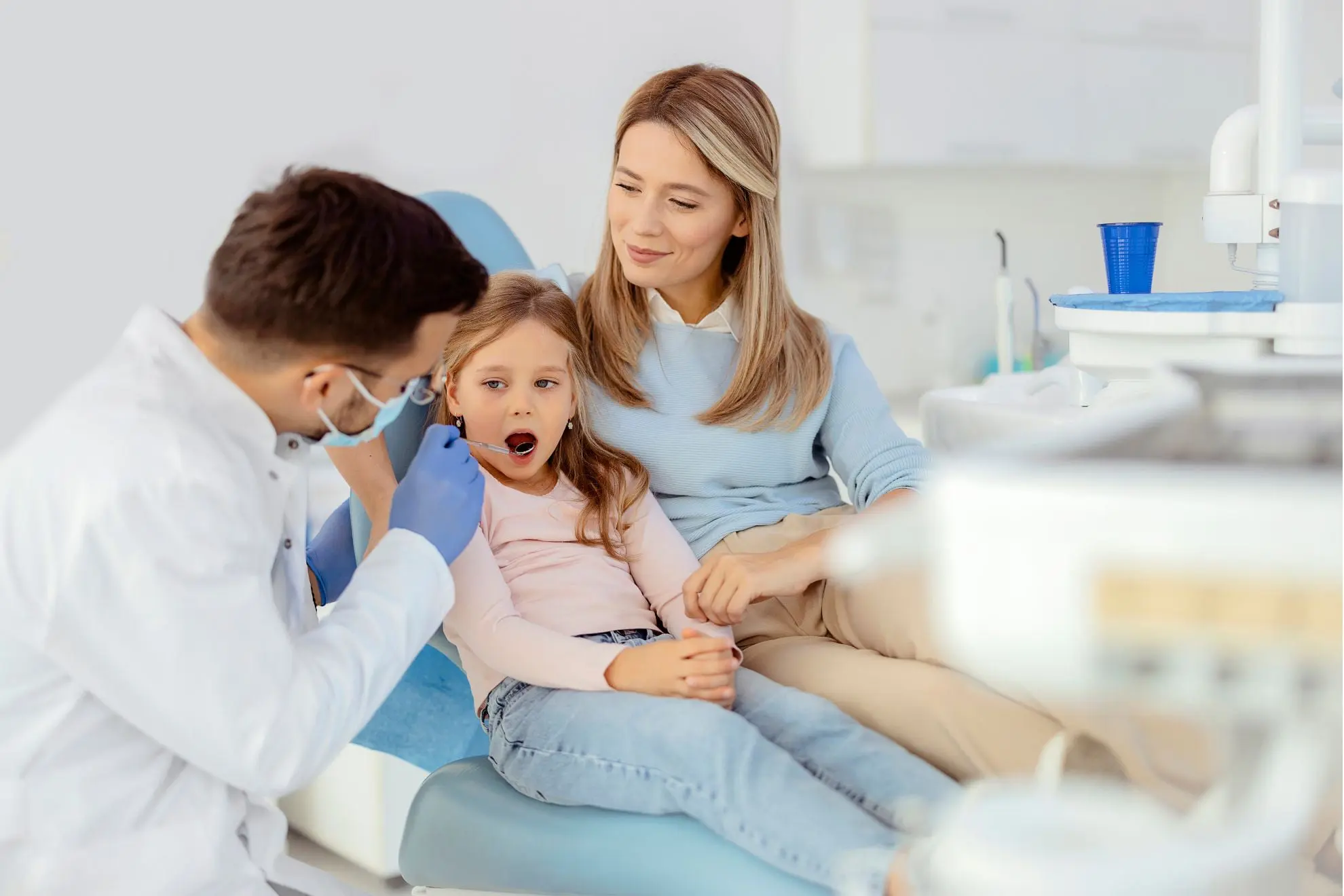 At Levine Orthodontics in Fort Myers, Cape Coral, or Lehigh Acres, FL, a masked dentist examines a child's mouth for signs of tongue thrust, with the child's mother by their side.