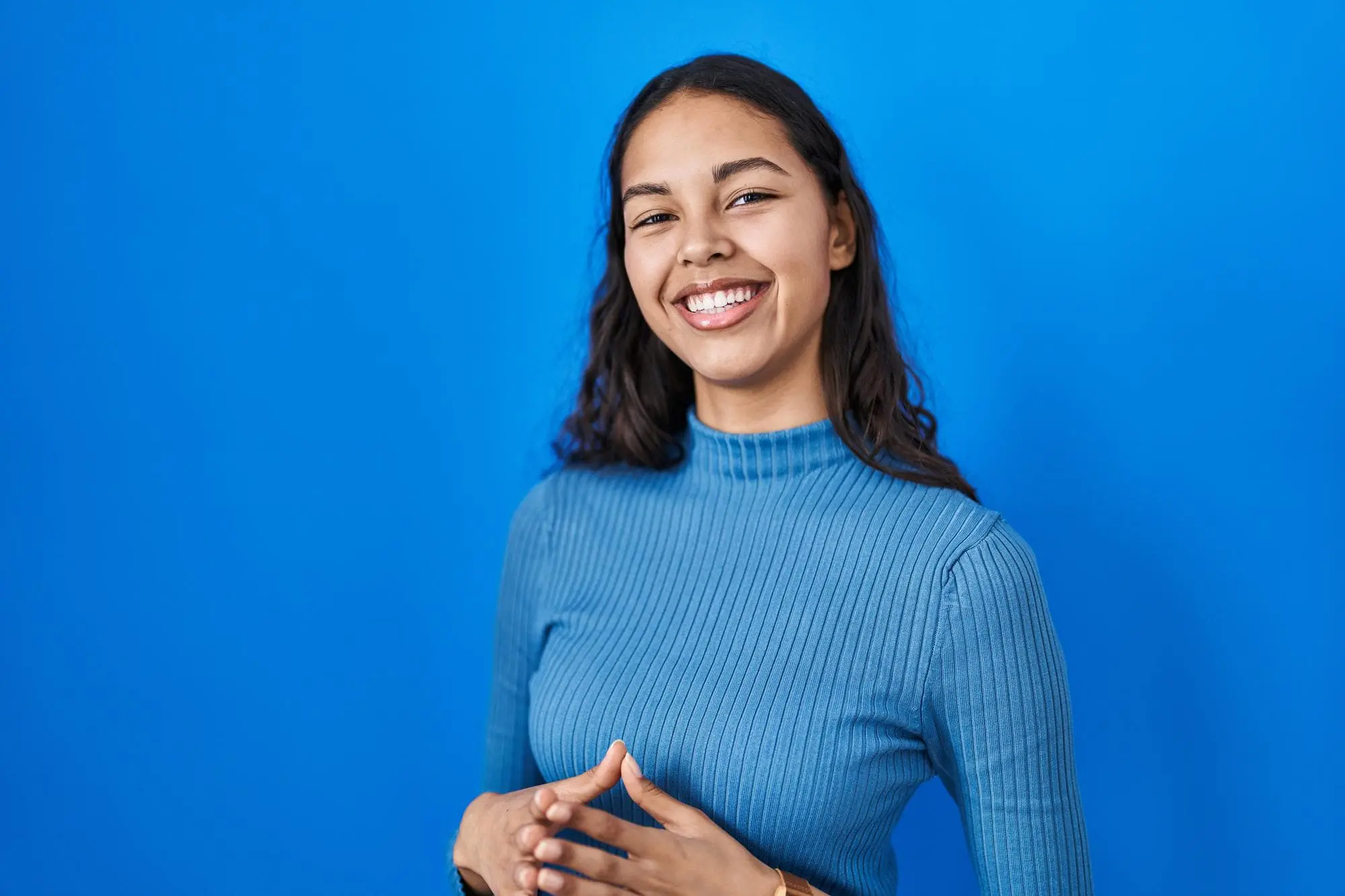 A woman with long dark hair, dressed in a blue ribbed turtleneck, smiles against a solid blue background at Levine Orthodontics in Fort Myers, Cape Coral or Lehigh Acres, FL.