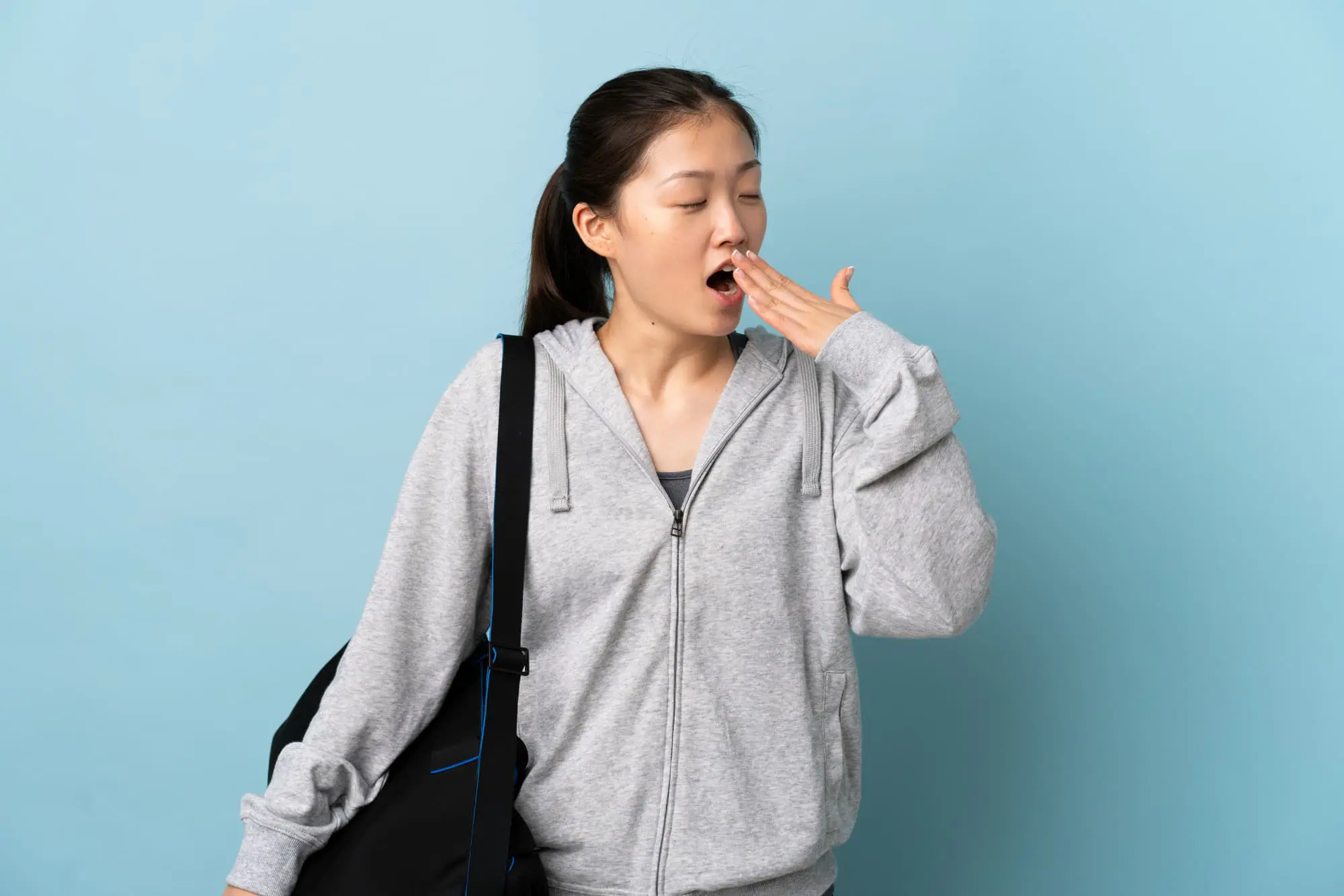 Yawning woman in gray hoodie with gym bag, light blue background; Levine Orthodontics in Cape Coral, FL highlights airway orthodontics for better sleep.
